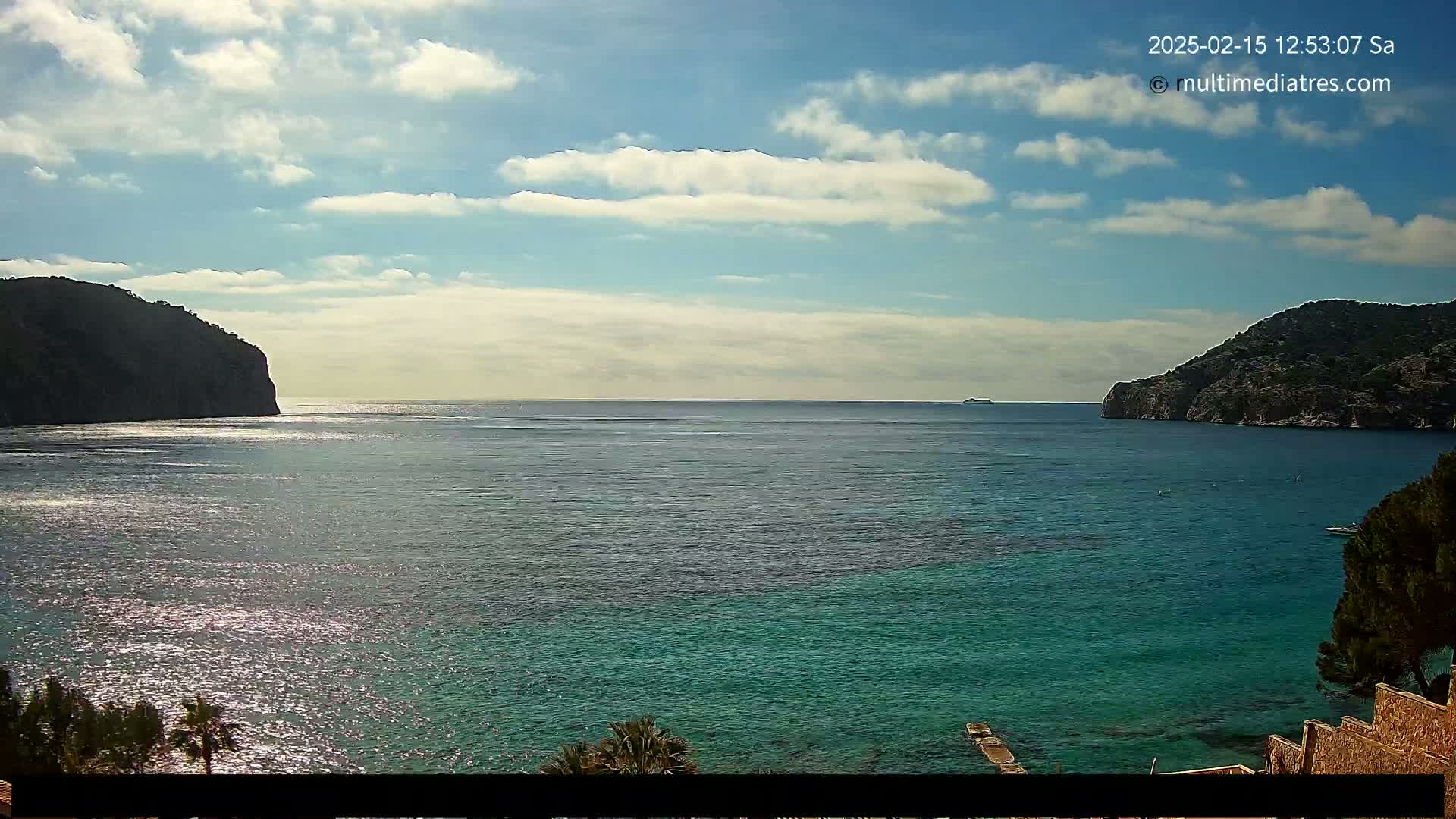 A partly sunny day shows a calm, teal-colored bay nestled between two dark, rocky hills, with a small boat and distant vessel visible on the water.