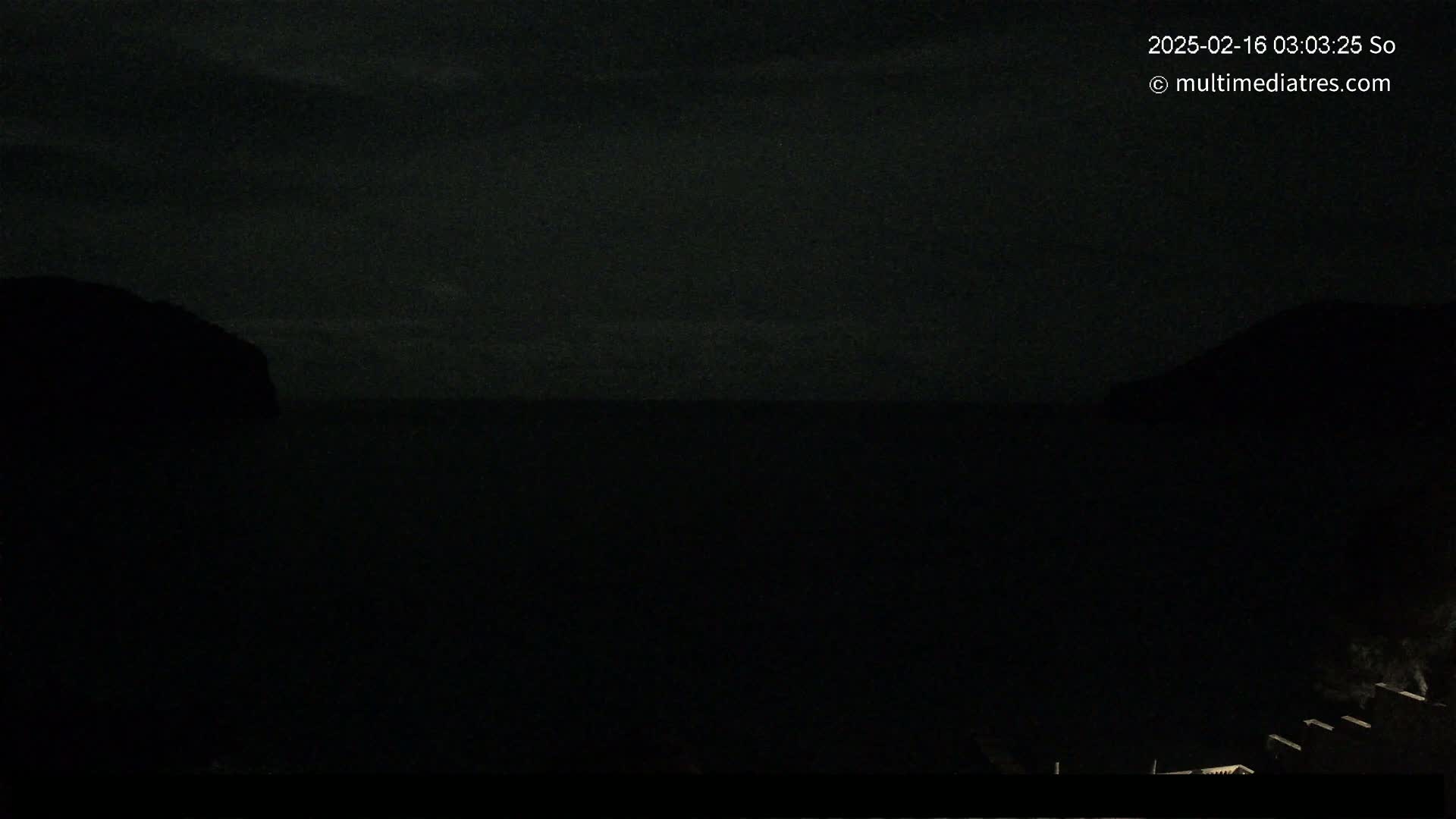 A dark nighttime coastal scene shows a mostly obscured body of water between two shadowy landmasses under a cloudy sky.