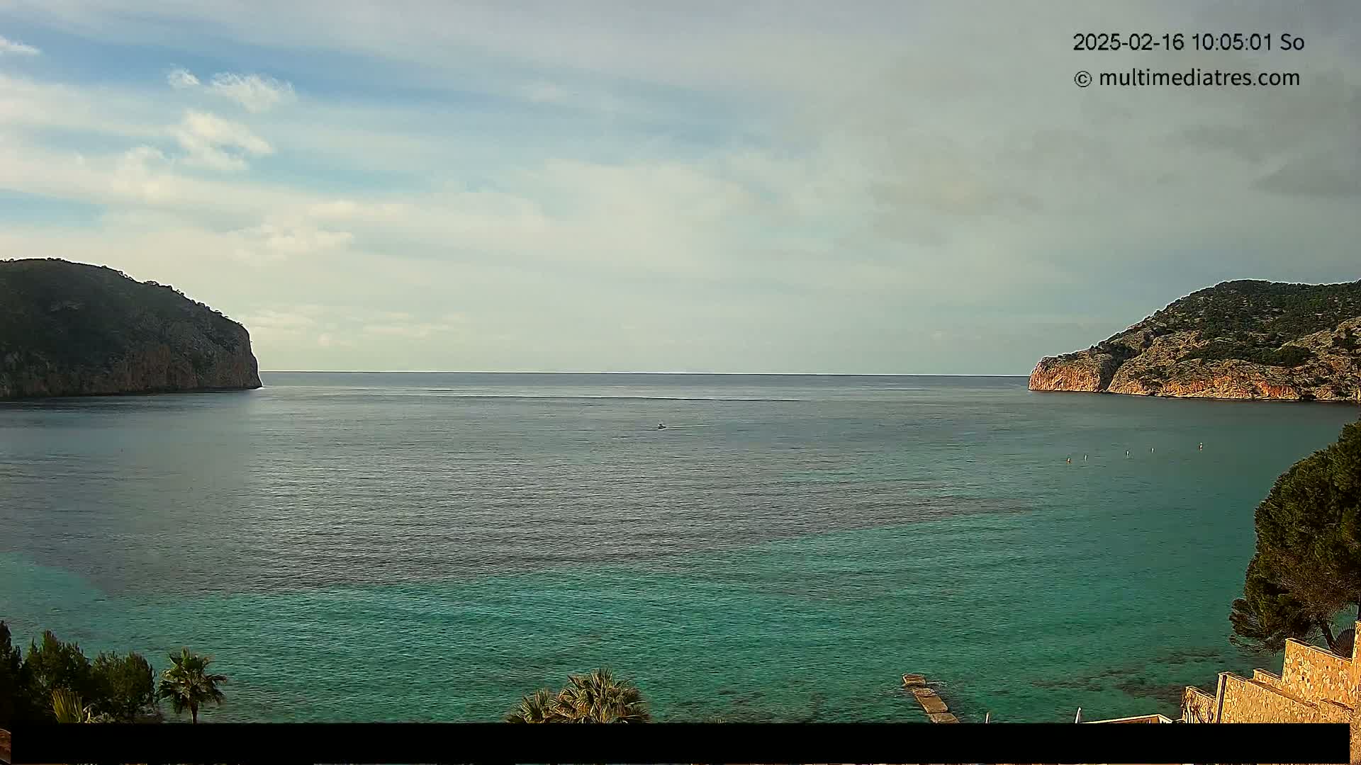 A calm, partly cloudy day reveals a picturesque coastal scene with two rocky hills flanking a teal-colored bay.