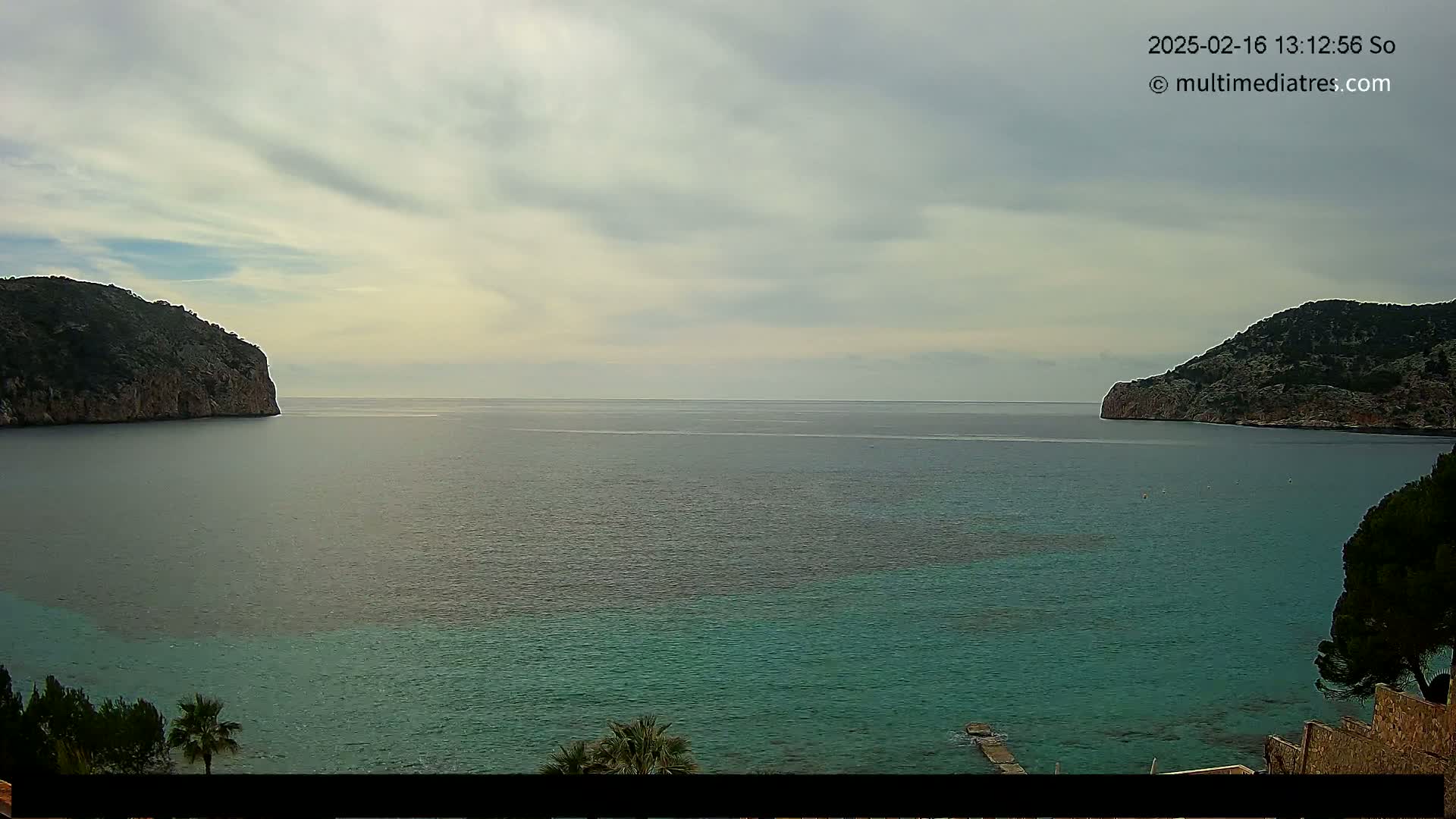 A calm, partially cloudy day shows a bay of teal water between two dark, rocky hillsides.