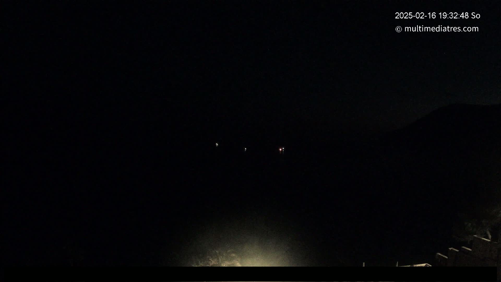 A dark nighttime scene shows a few distant lights and a faint hillside under a clear, dark sky.