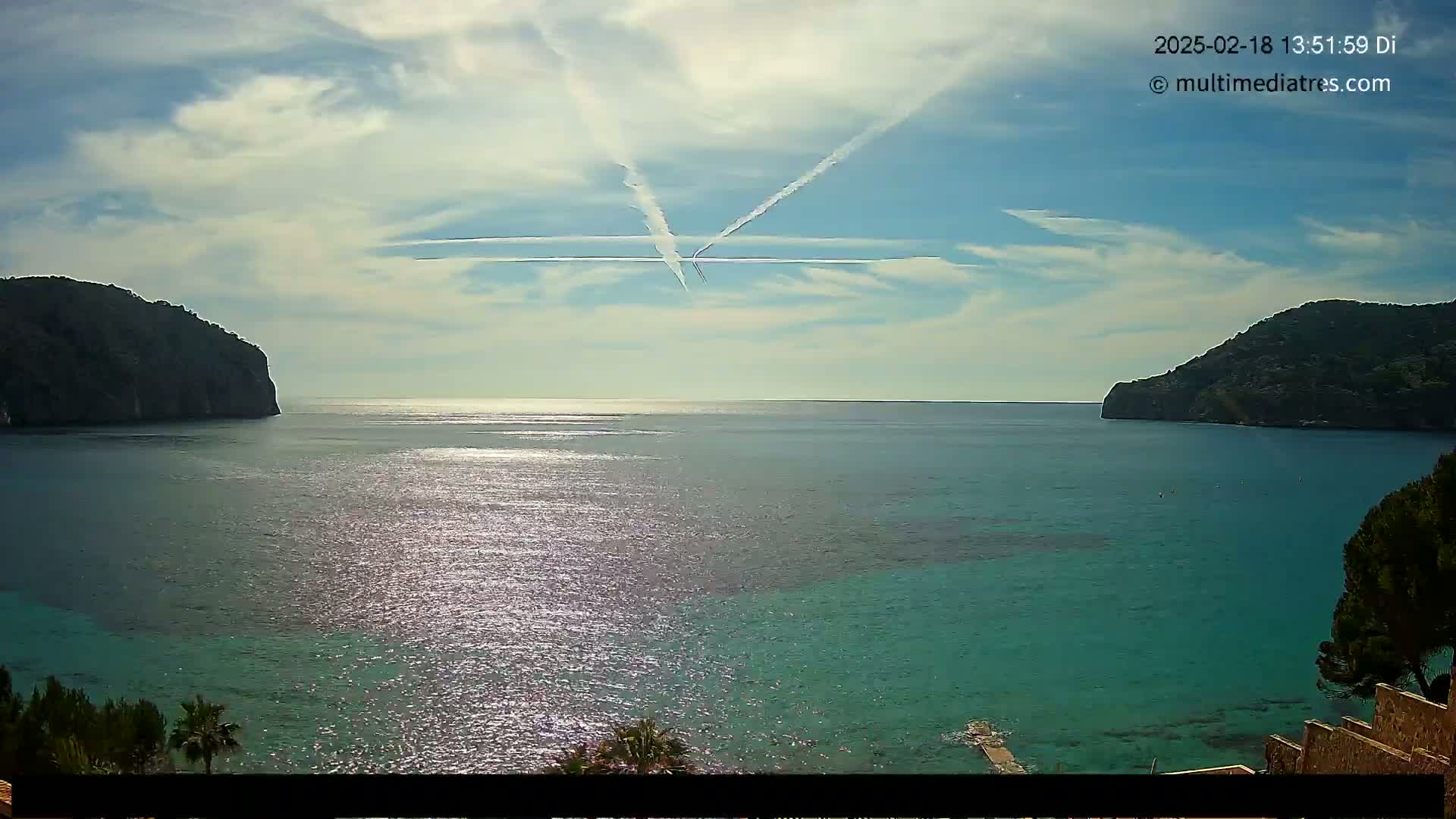 A partly cloudy day shows two dark green hills flanking a calm, teal ocean with contrails crossing the sky.