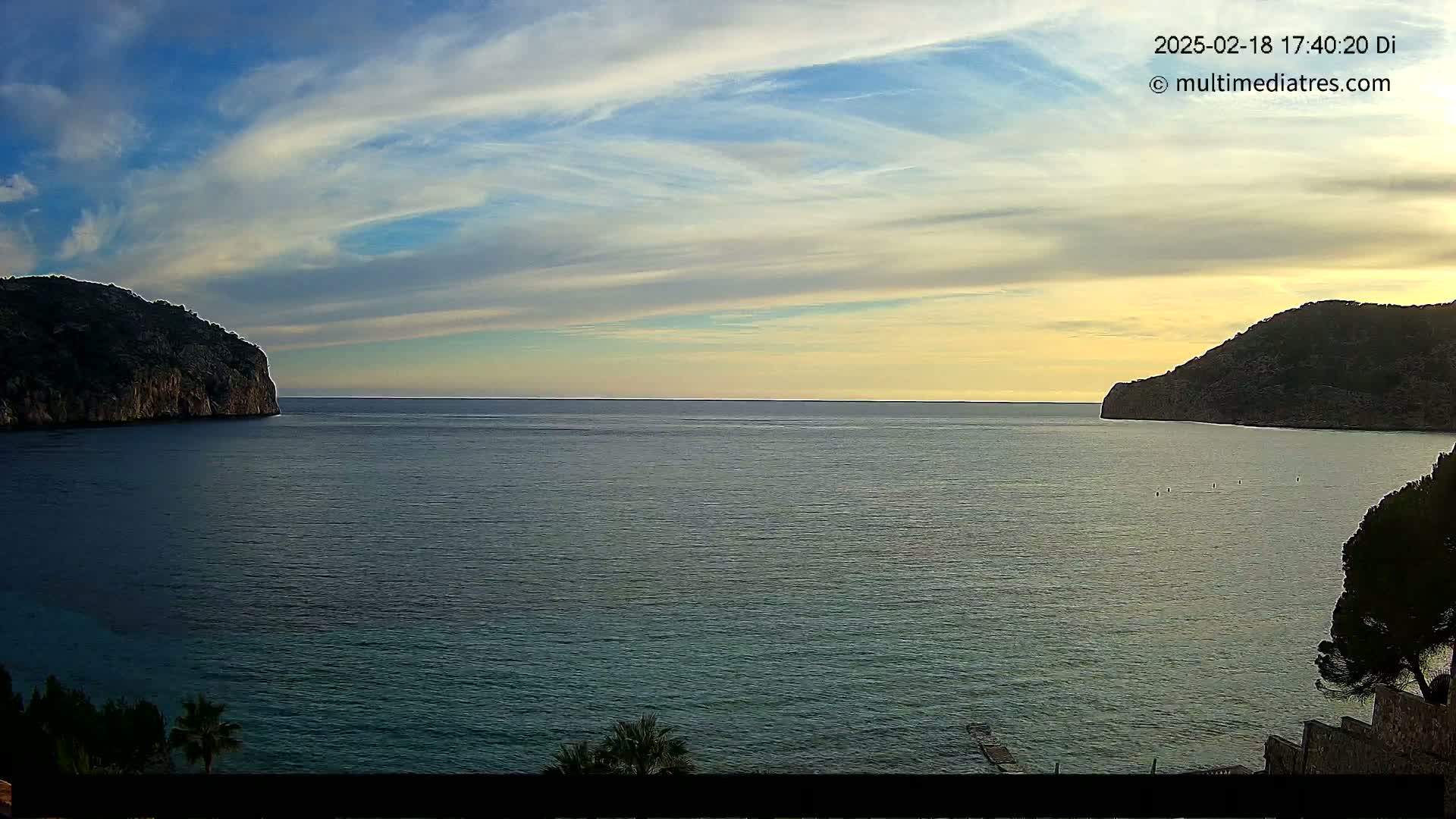 A calm sea is nestled between two dark, rocky hills under a partly cloudy, sunset sky.
