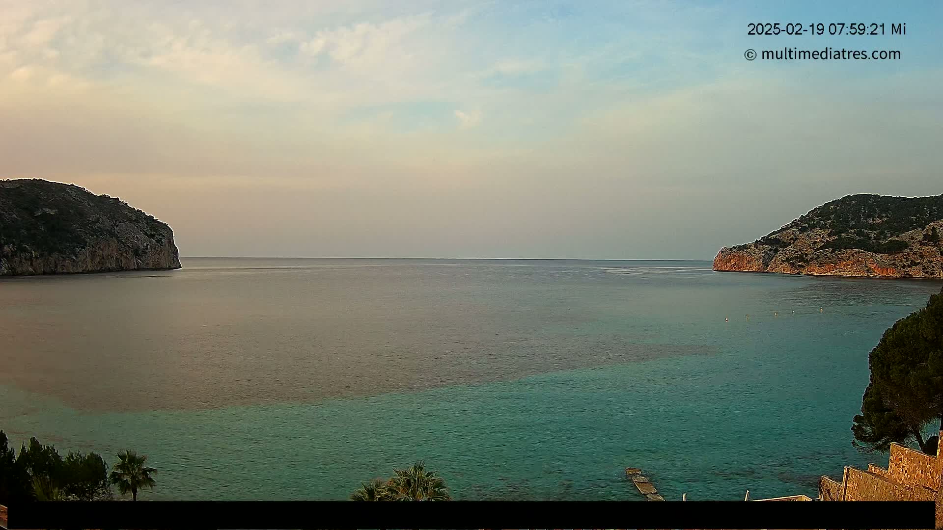 A calm, pale-blue sea is nestled between two rocky cliffs under a partly cloudy sky.