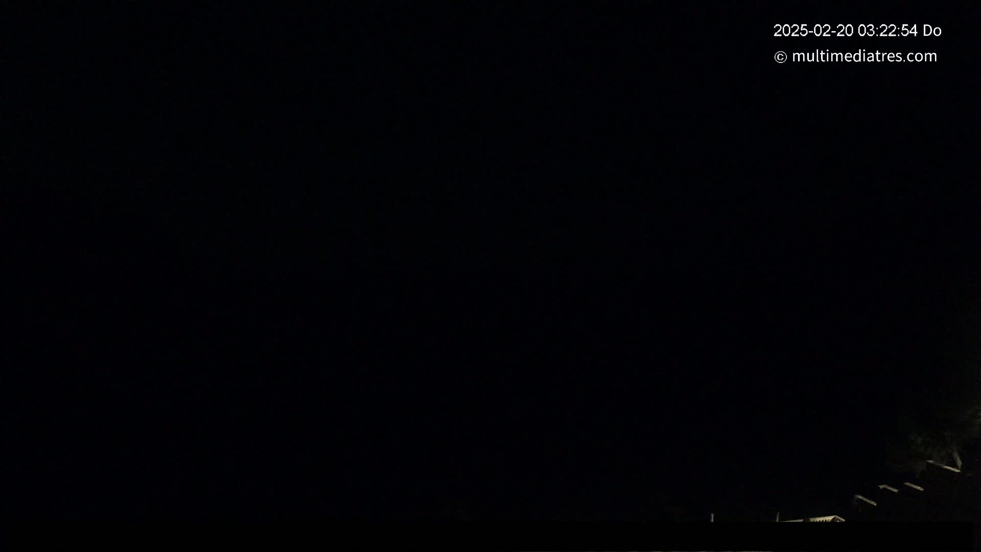 A dark nighttime scene shows a few dimly lit structures at the bottom of the frame against a completely black sky.
