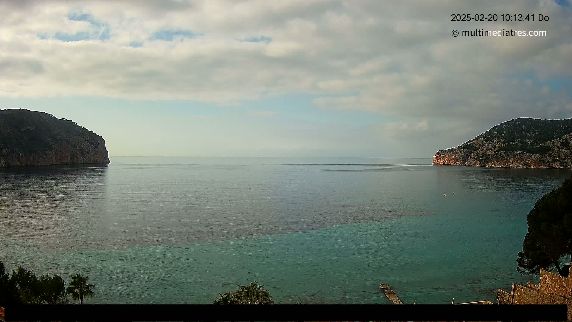 A partly cloudy day reveals a calm sea separating two rocky headlands, with a small jetty visible near the lower right.