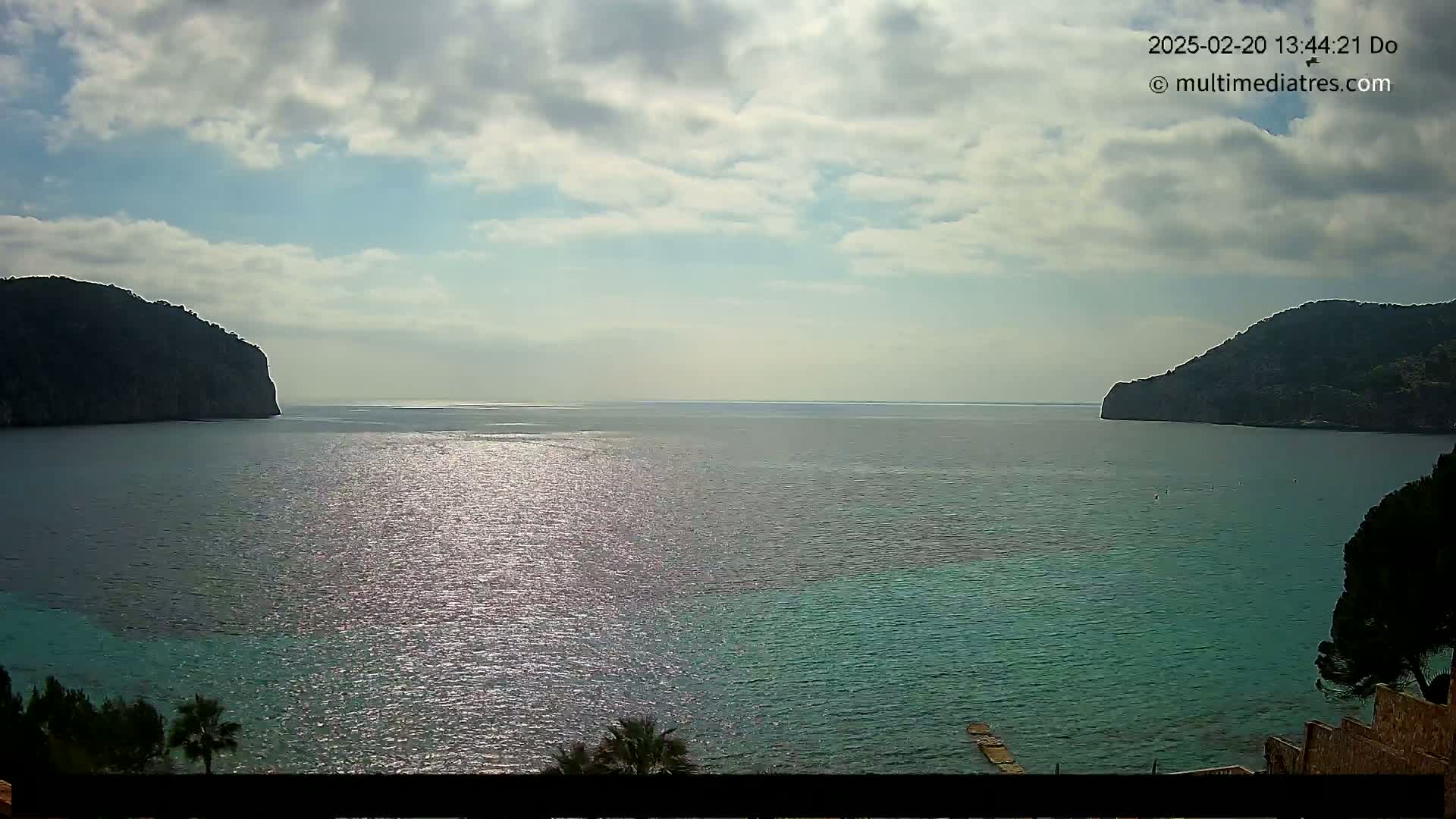 A partly cloudy day shows a calm, teal sea separating two dark, rocky cliffs under a mostly cloudy sky.