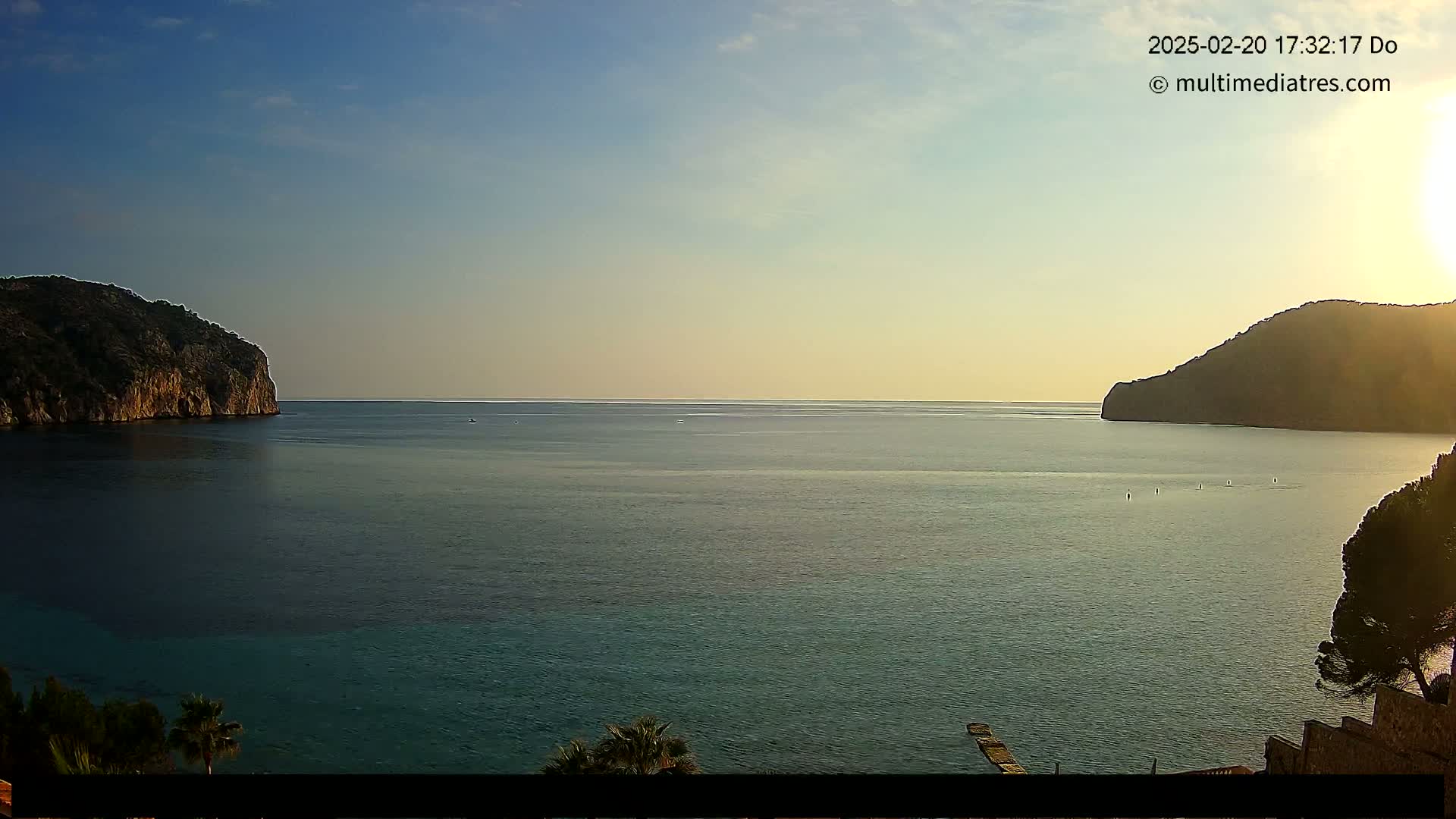 A calm sea at sunset is nestled between two rocky hills, with a few people visible in the distance.