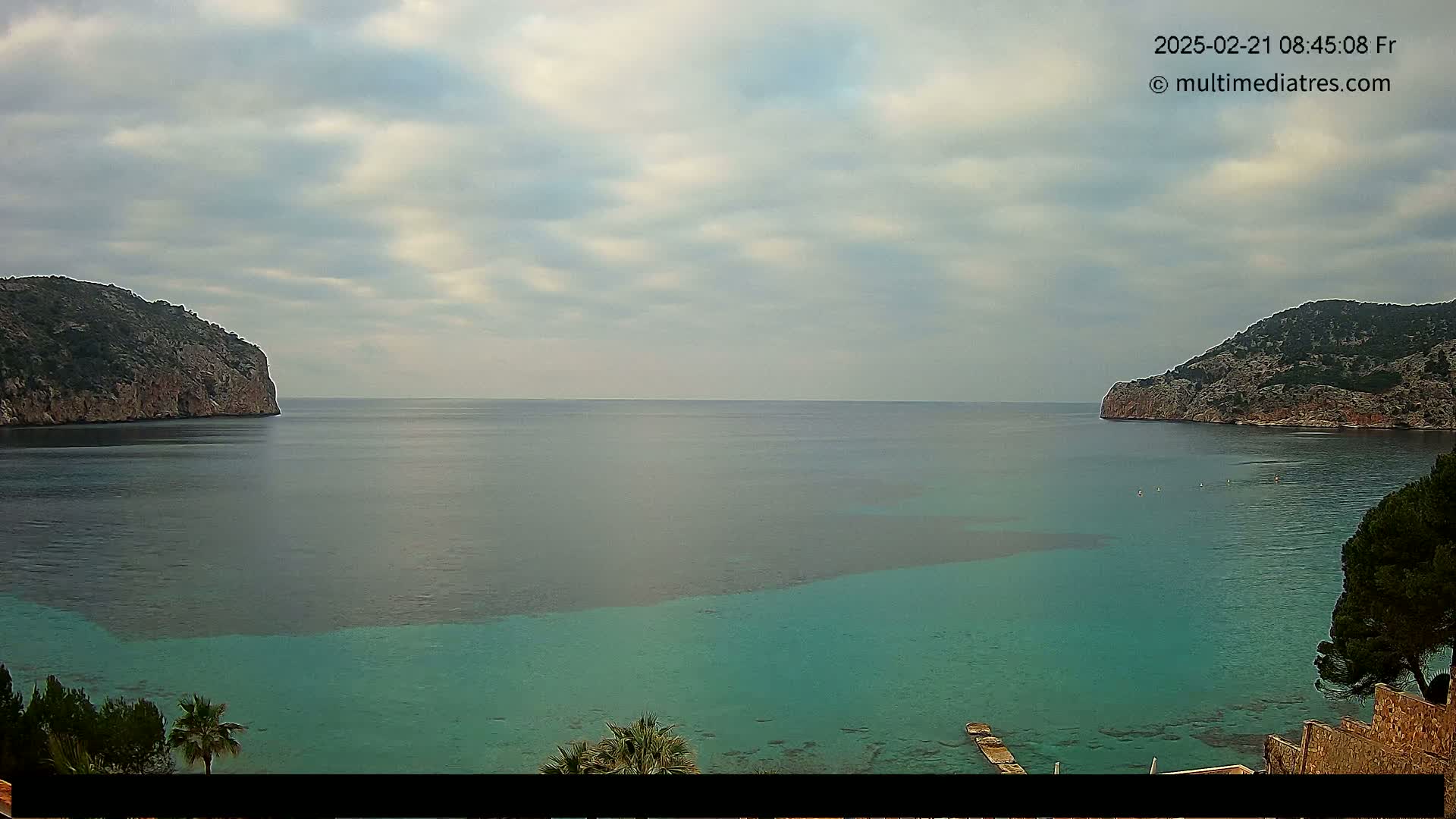 A tranquil bay with teal-colored water is nestled between two rocky hills under a cloudy sky.