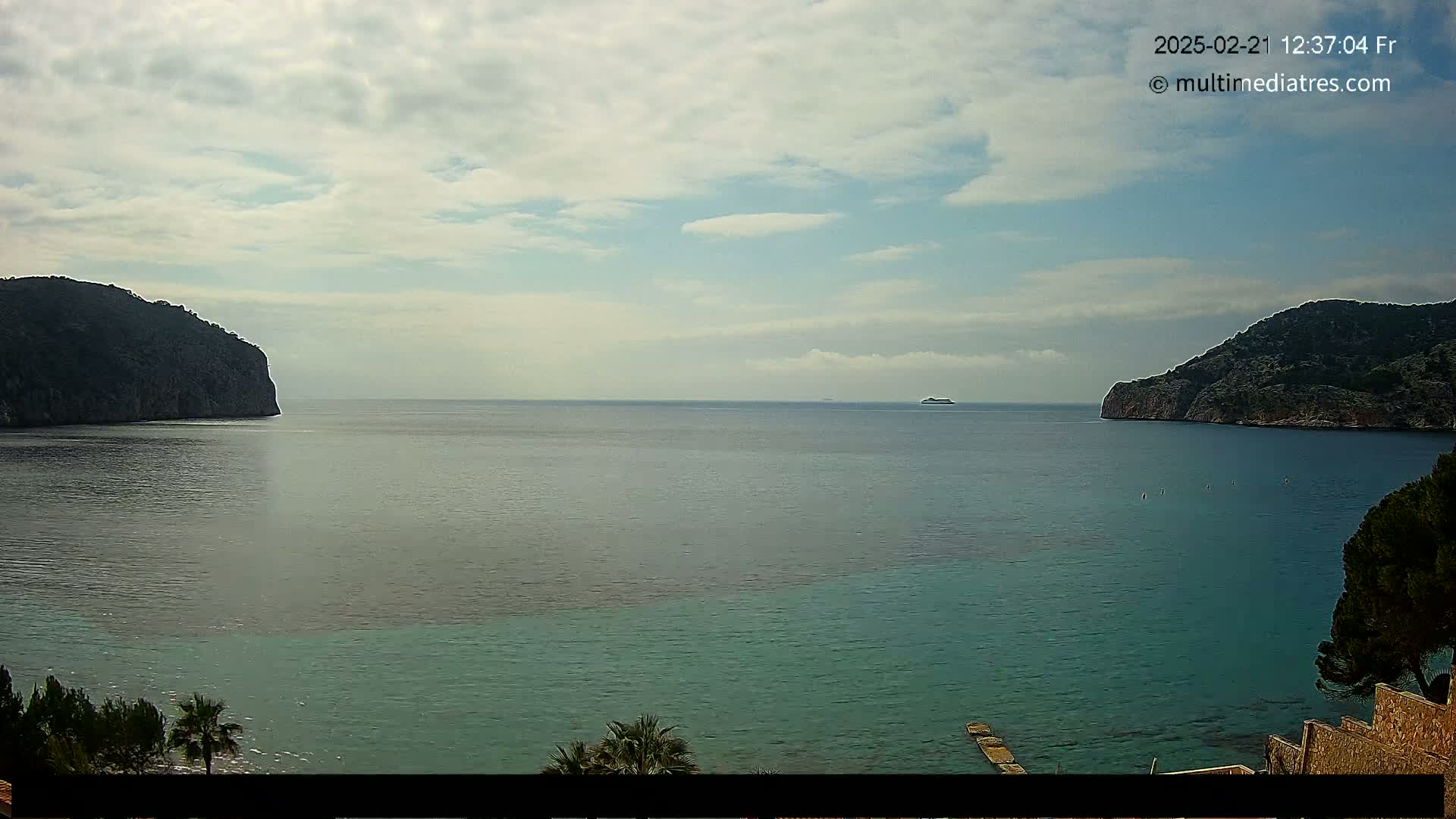 A partly cloudy day shows a calm sea with two dark, rocky headlands framing a view of a distant ship.