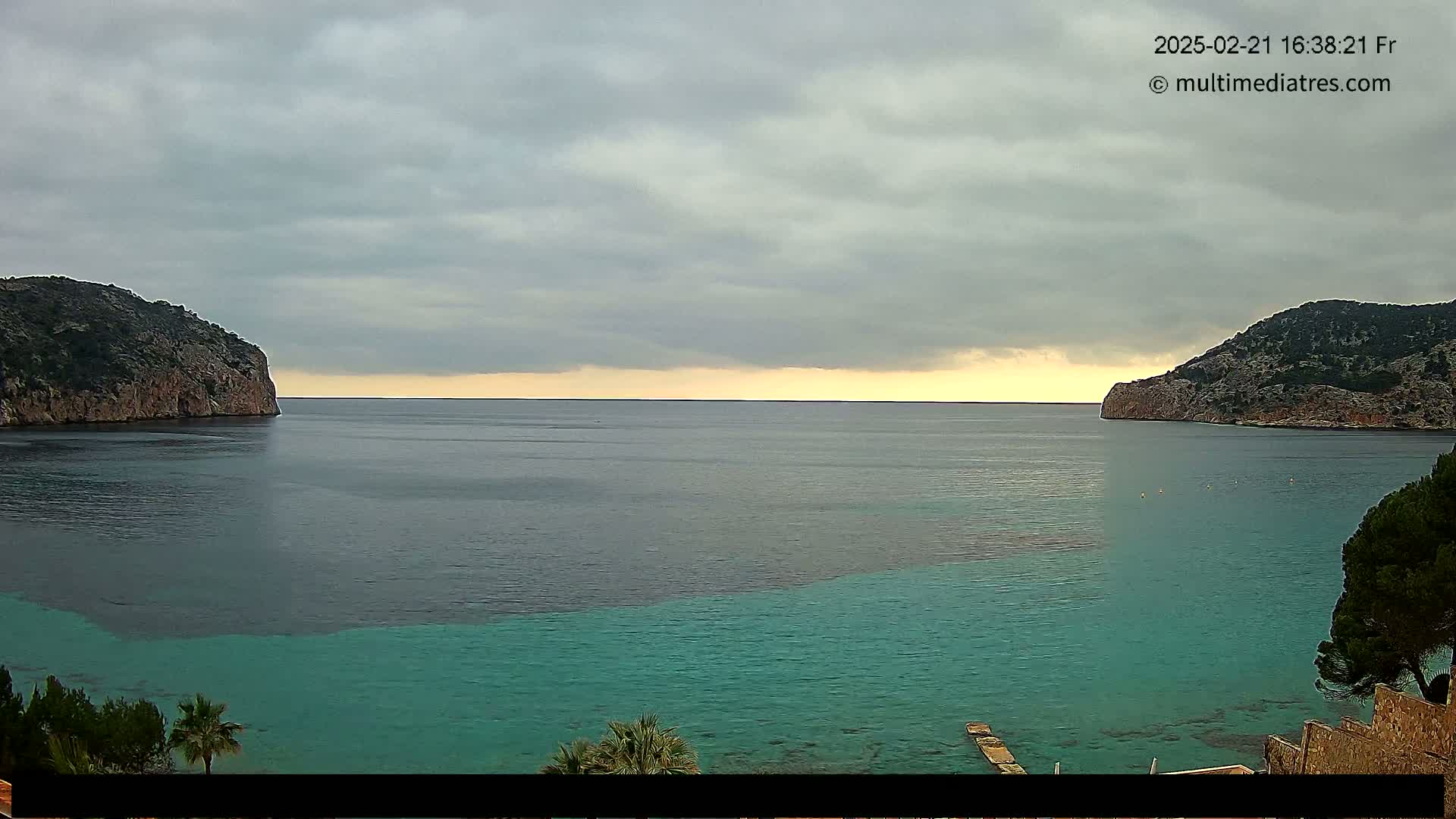 A calm bay with teal water is nestled between two rocky hills under a cloudy sky.