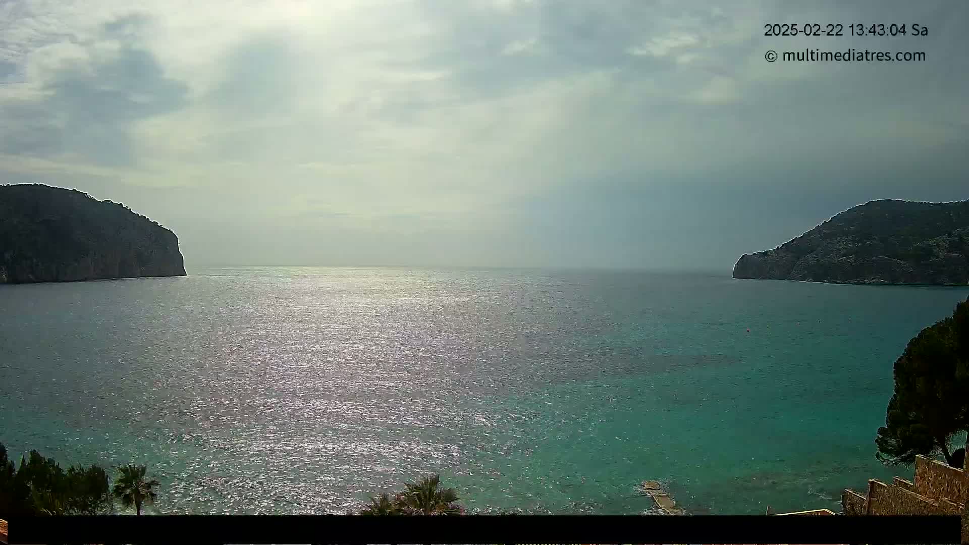 A partly cloudy sky overlooks a calm, shimmering sea separating two dark, rocky hillsides.