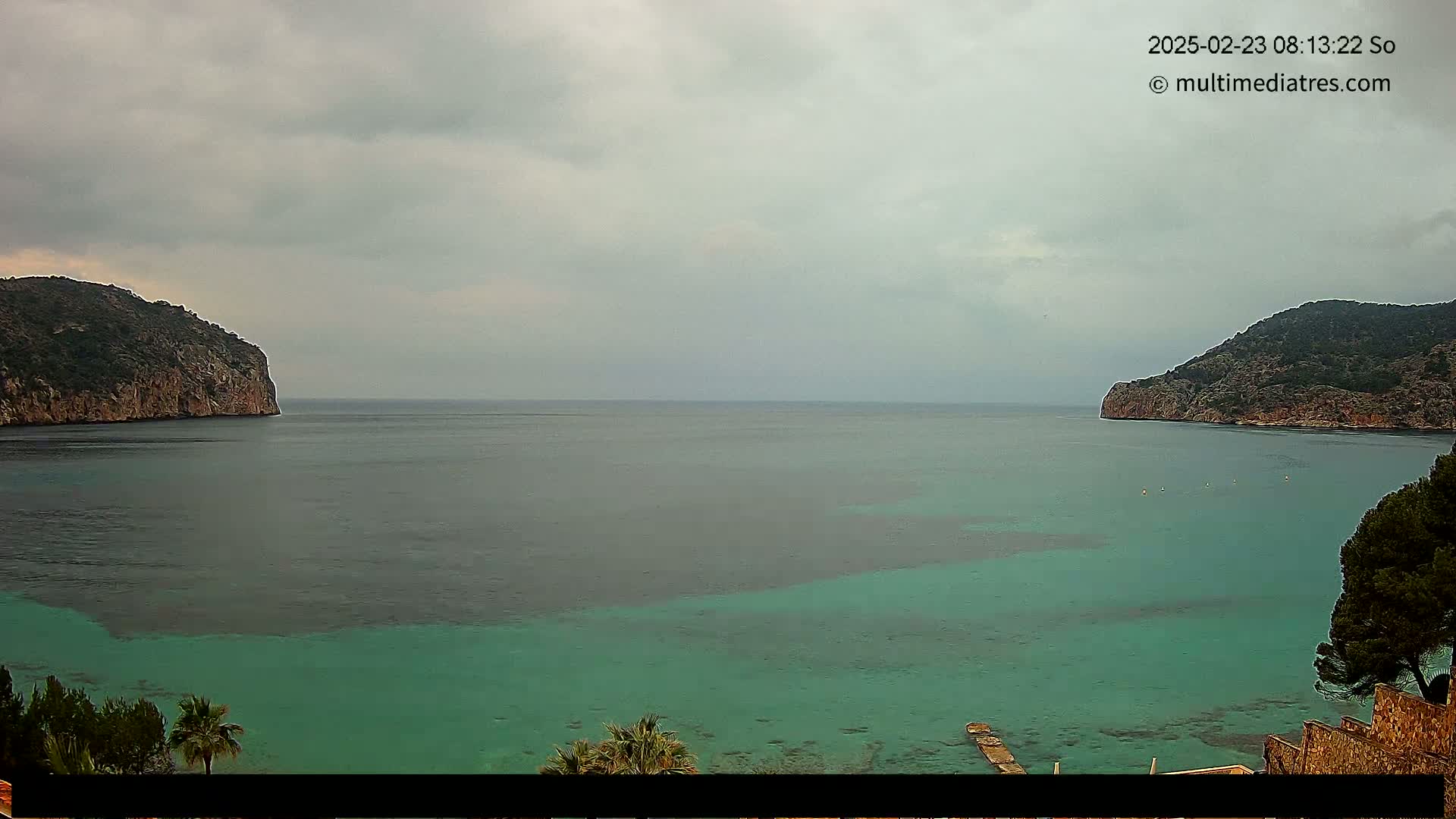 A calm, teal-colored bay is nestled between two rocky hills under an overcast sky.