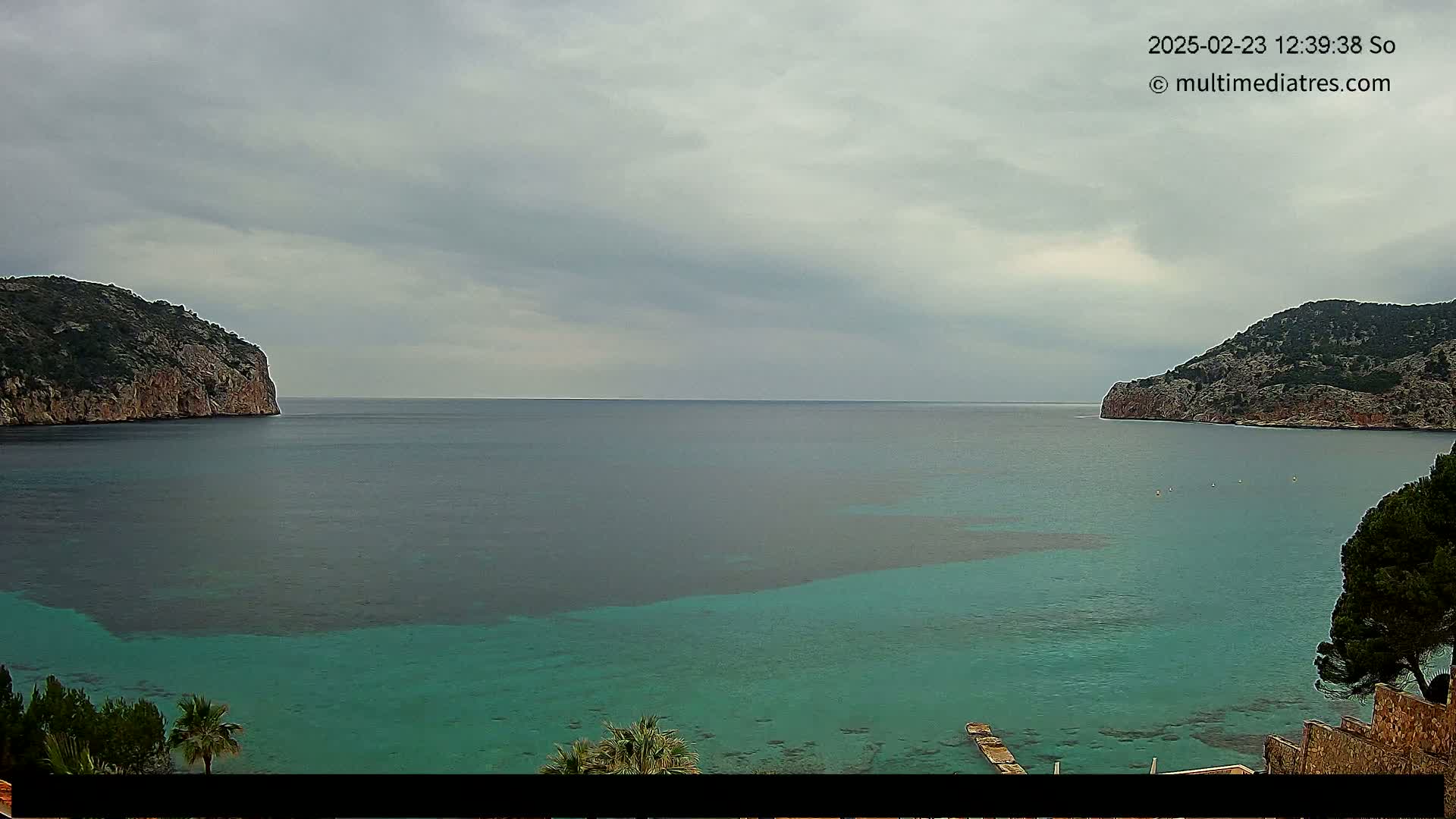 A calm, teal-colored bay is nestled between two rocky hills under an overcast sky.