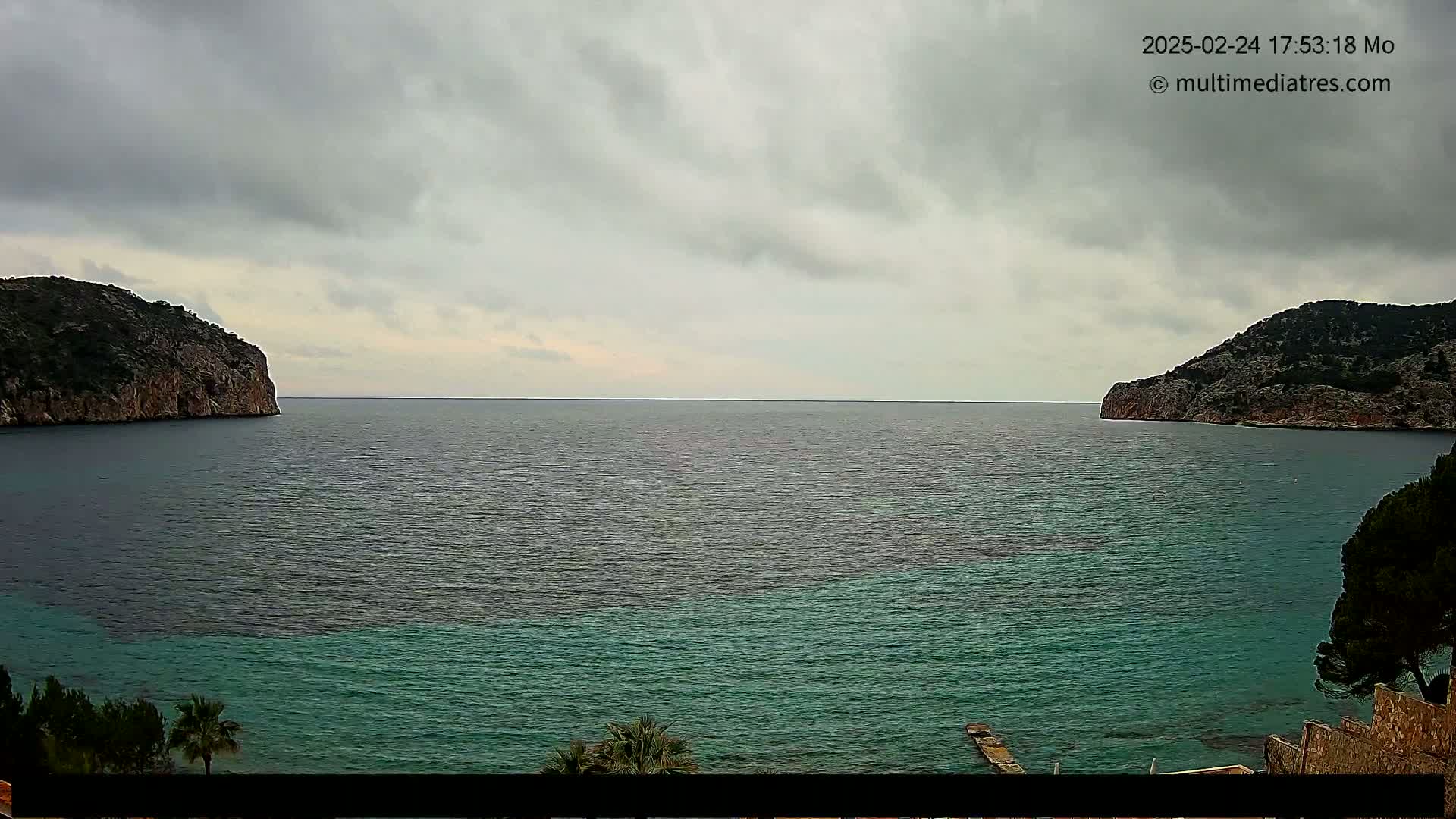 A calm, overcast day shows a view of a teal-colored bay between two rocky hillsides.