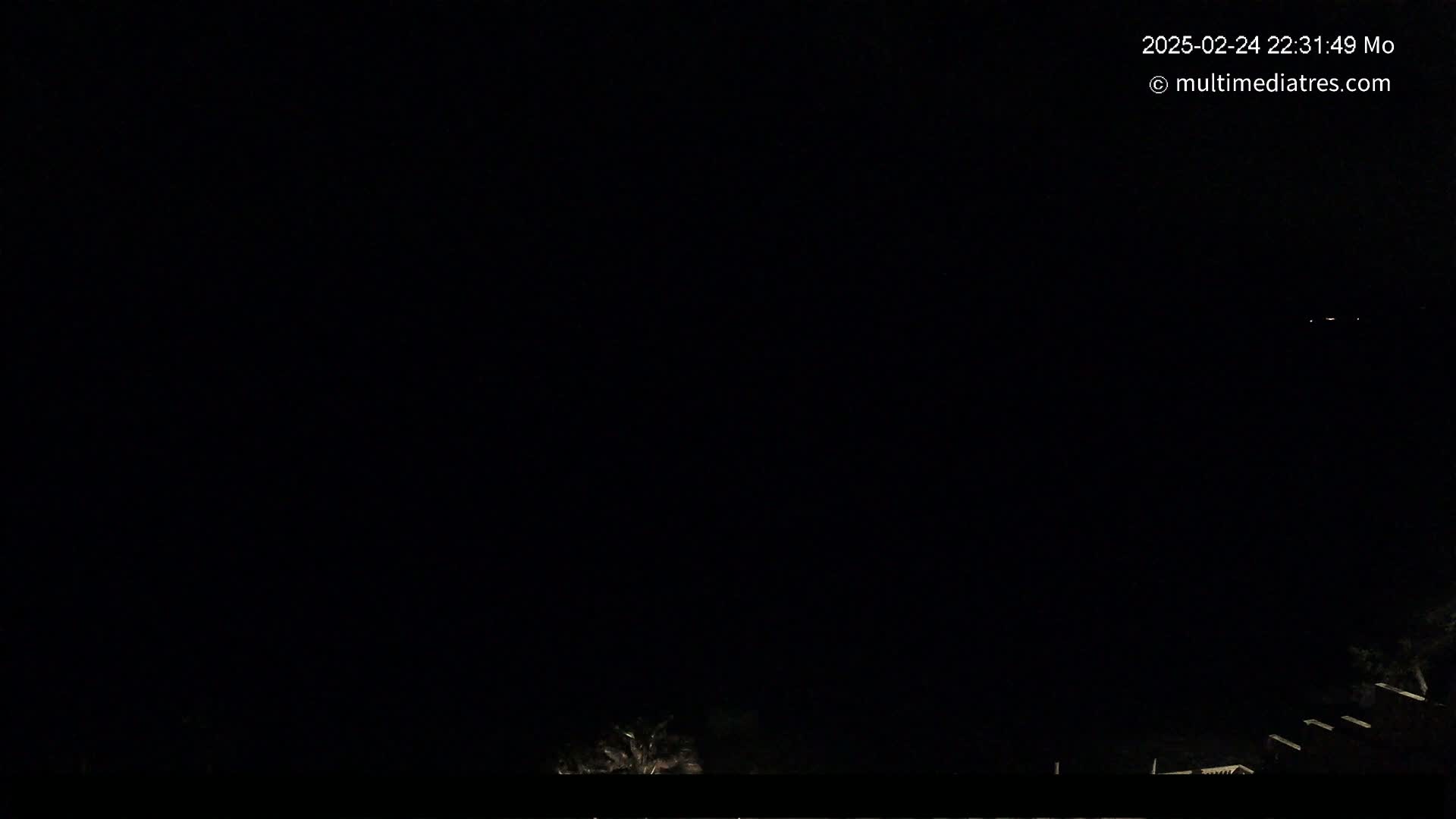 The image shows a dark night scene with a few faint lights visible in the distance and some dark vegetation at the bottom.