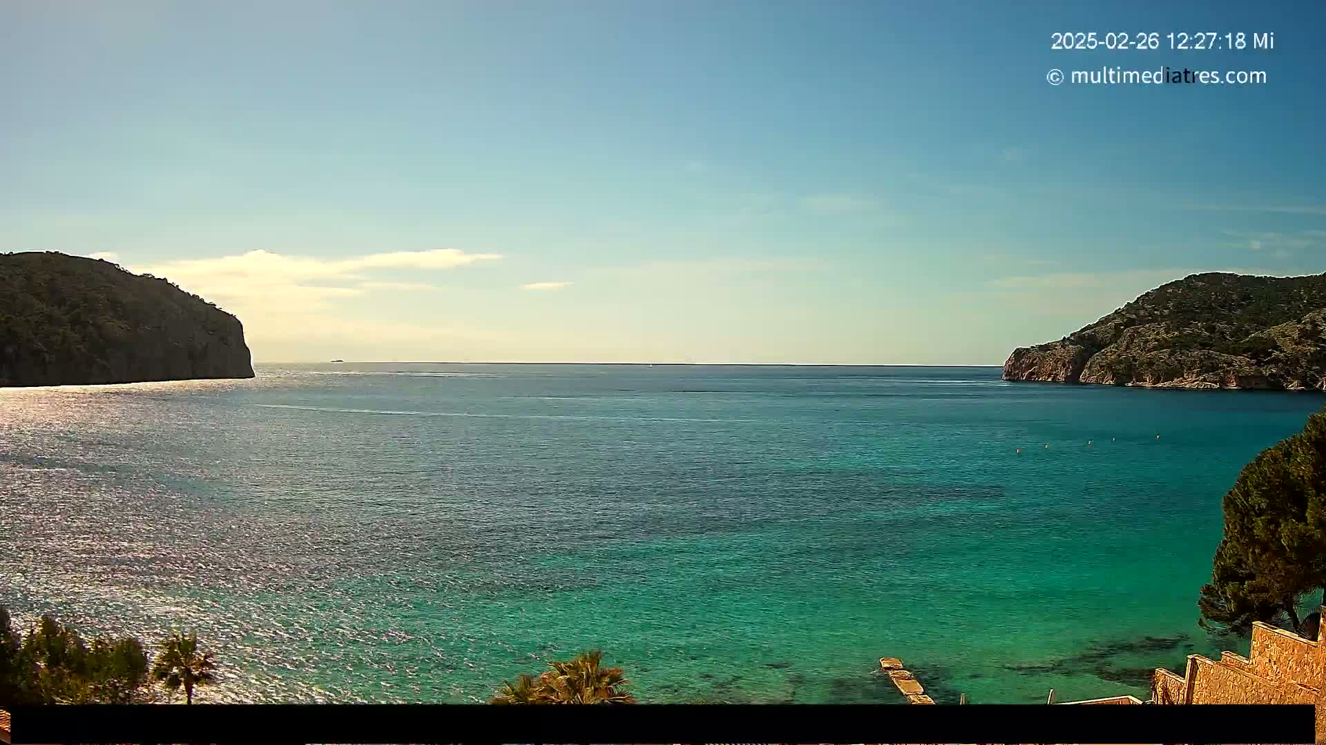 A tranquil, clear-blue ocean cove is nestled between two rocky hills under a mostly sunny sky with a few scattered clouds.