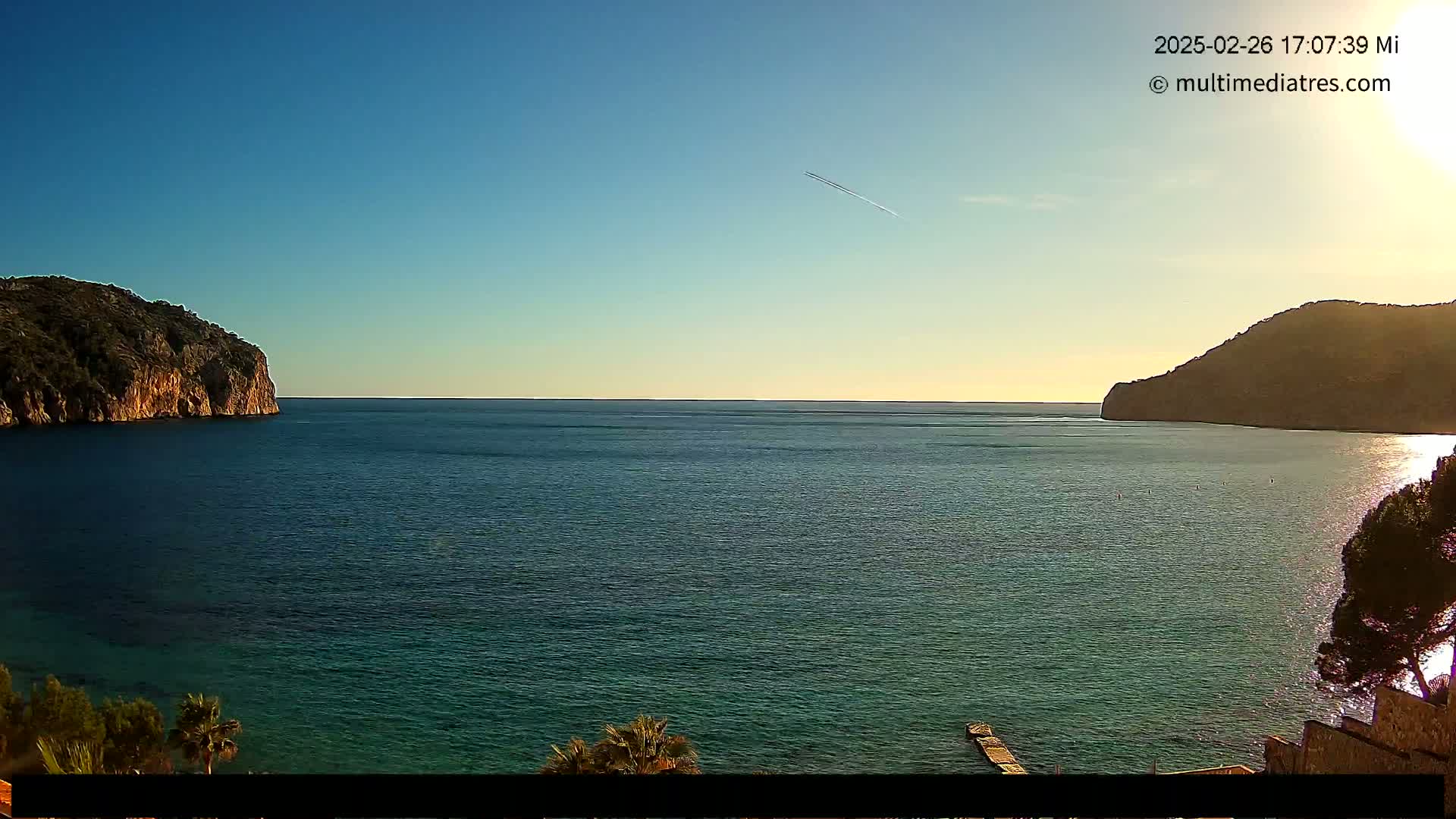 A tranquil sea scene shows calm, dark-blue water between two rocky hills under a clear, sunny sky with a single contrail visible.
