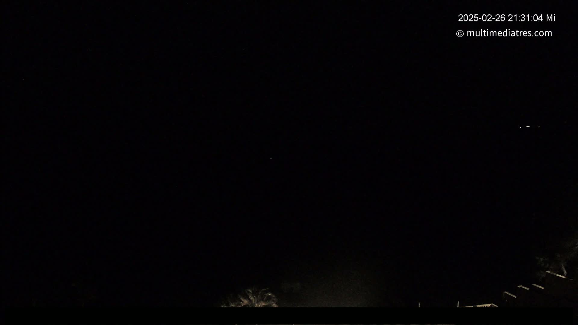 A nighttime outdoor scene shows a dark sky with a few faint lights in the distance, and the tops of some trees and structures are visible at the bottom.