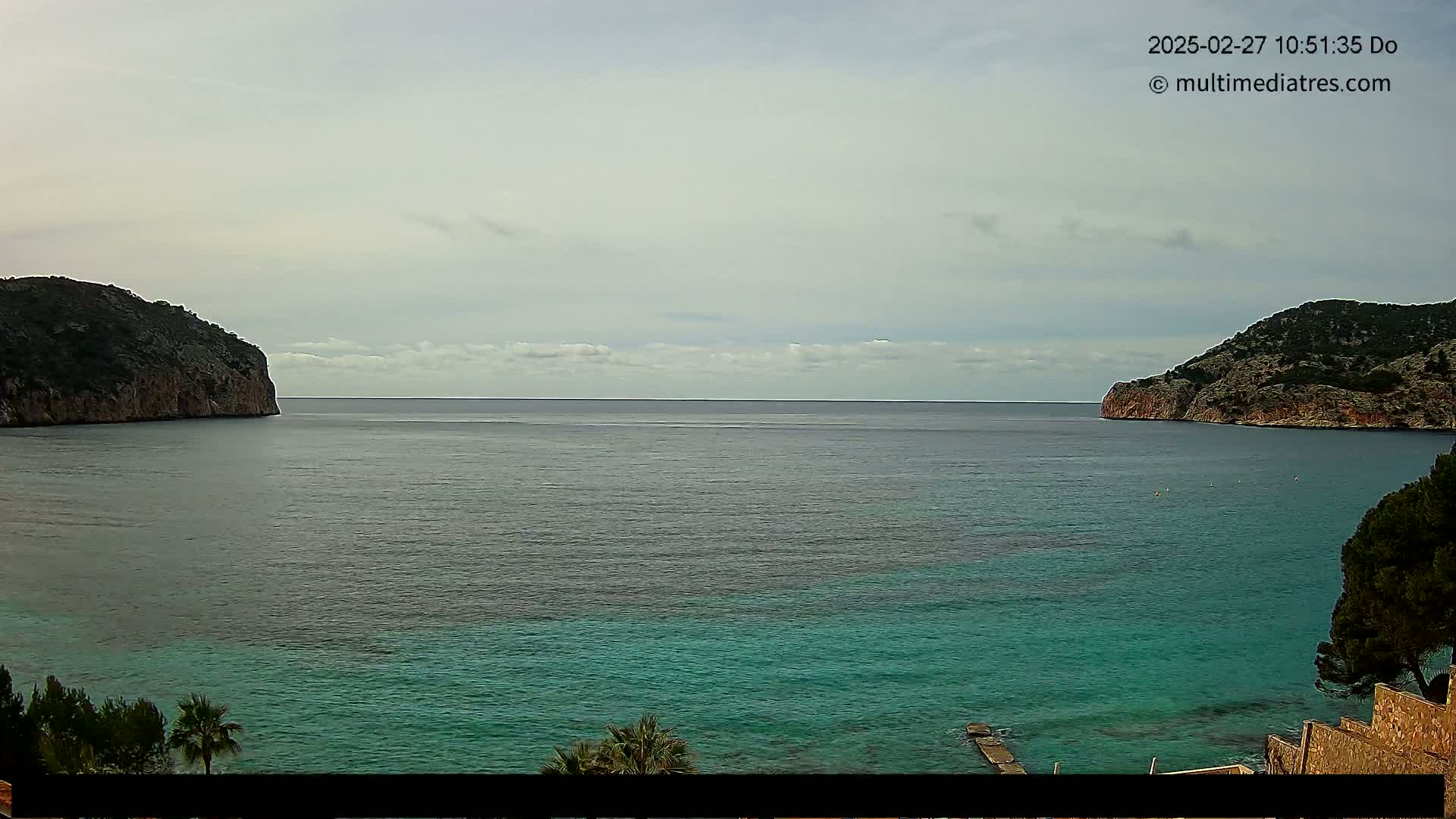 A tranquil bay with calm, teal water is nestled between two rocky headlands under a mostly cloudy sky.