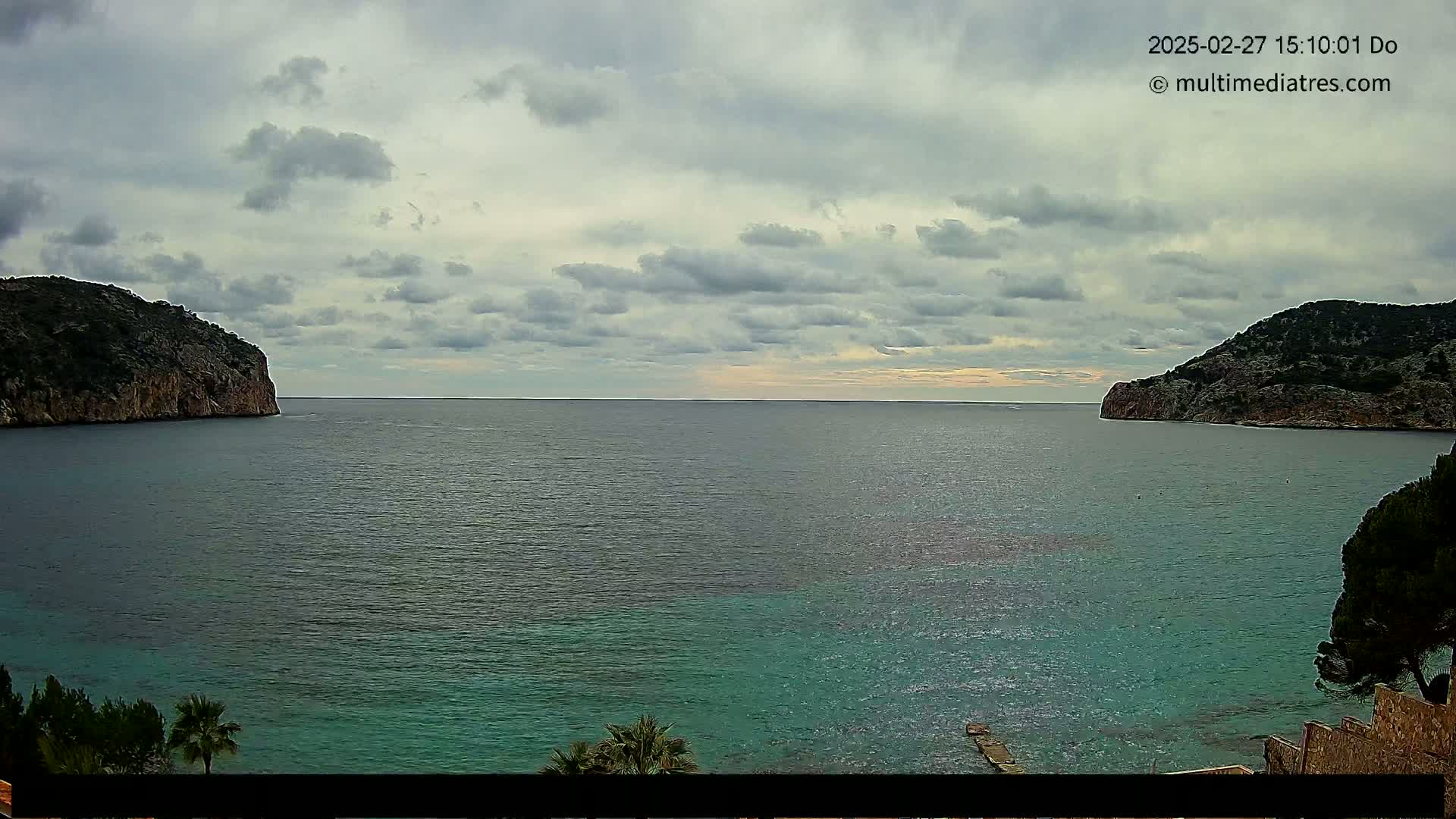 A calm, overcast day reveals a tranquil bay nestled between two rocky hillsides, the water a gradient of dark and teal.