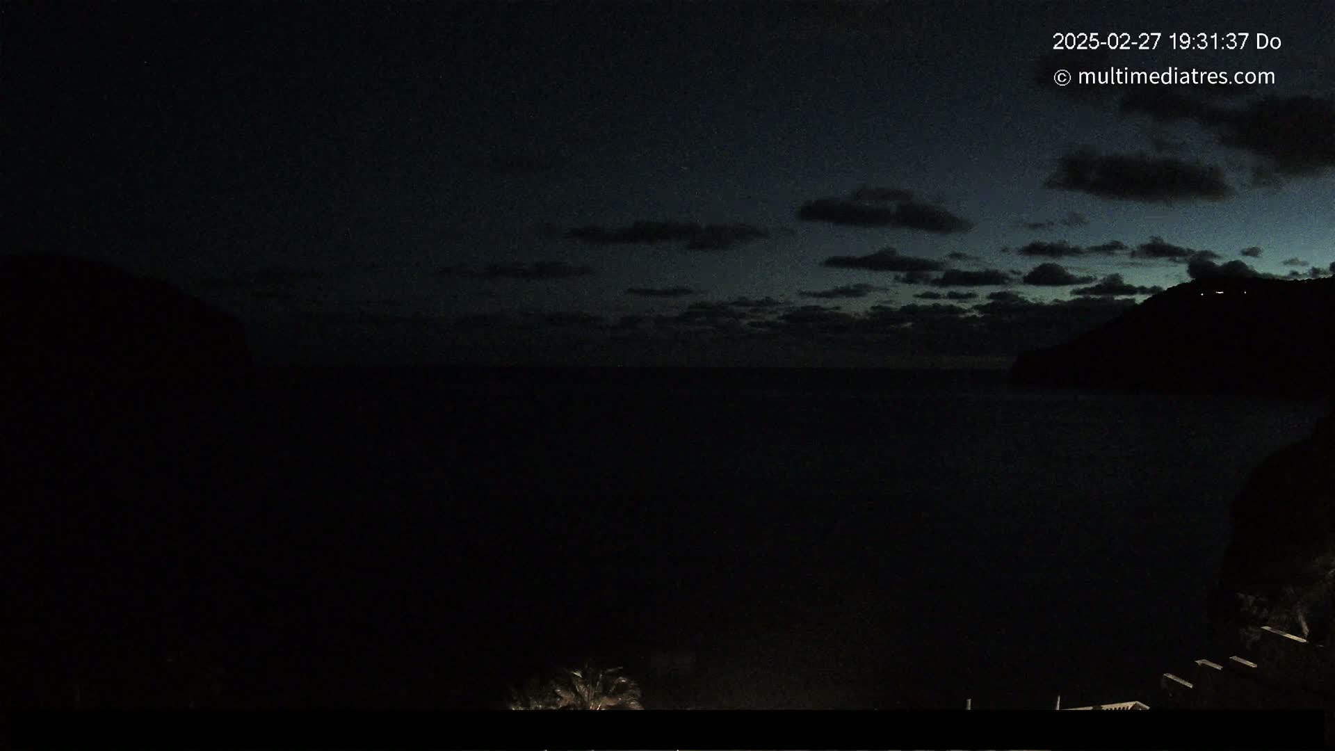 A dark, nighttime coastal scene shows a mostly calm body of water between two silhouetted hills under a cloudy sky.