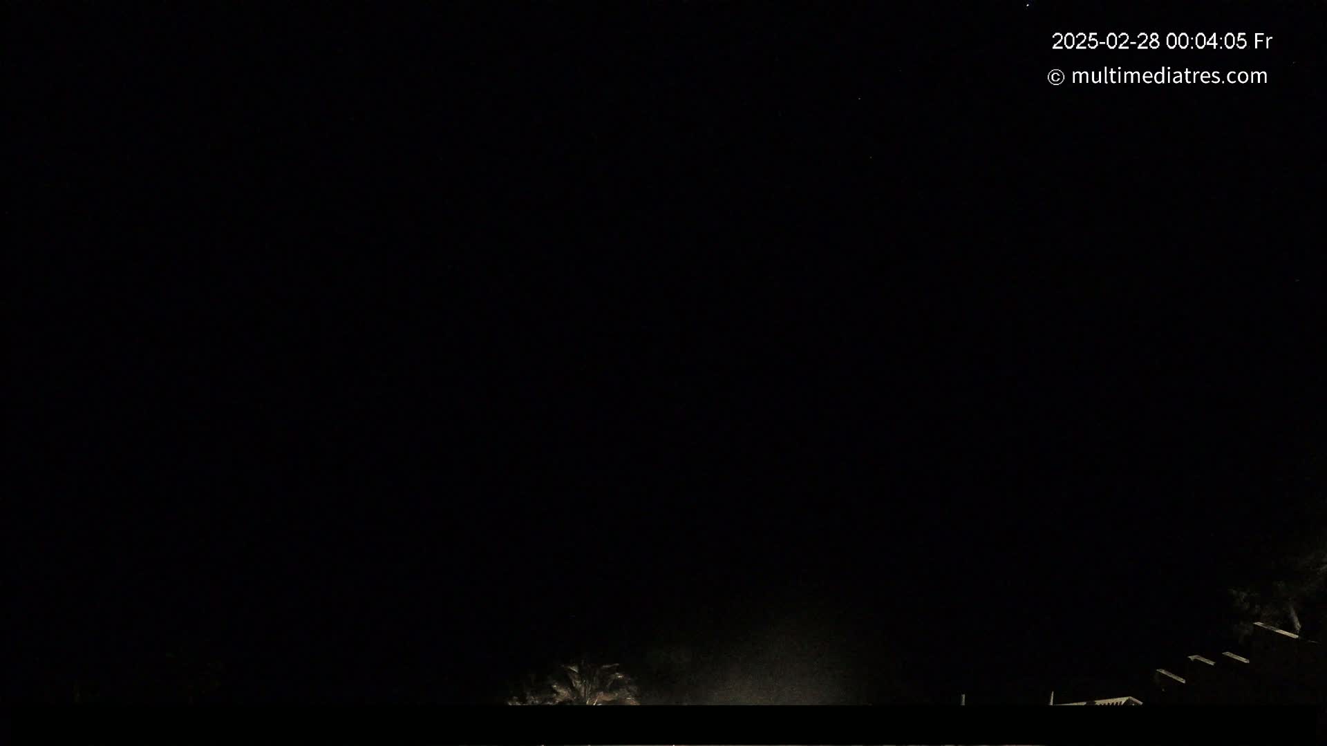 A nighttime outdoor scene shows a mostly dark sky with some faint light near the bottom and a few dark silhouettes of trees or structures.