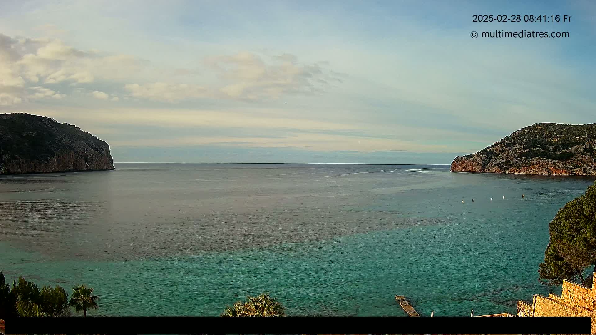 A calm sea with a teal-colored near shore and grayish-green farther out, is nestled between two rocky cliffs under a partly cloudy sky.