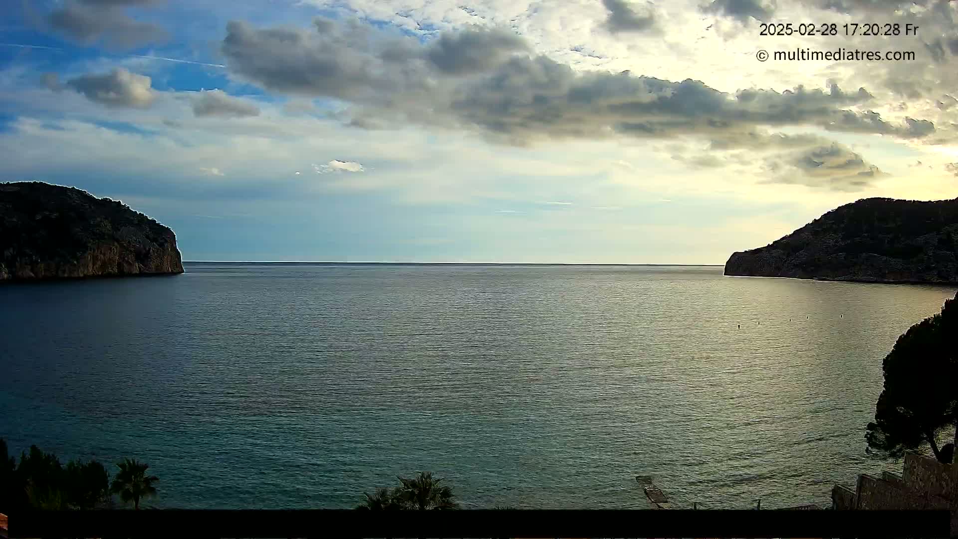A calm sea is nestled between two dark, rocky hills under a partly cloudy sky.