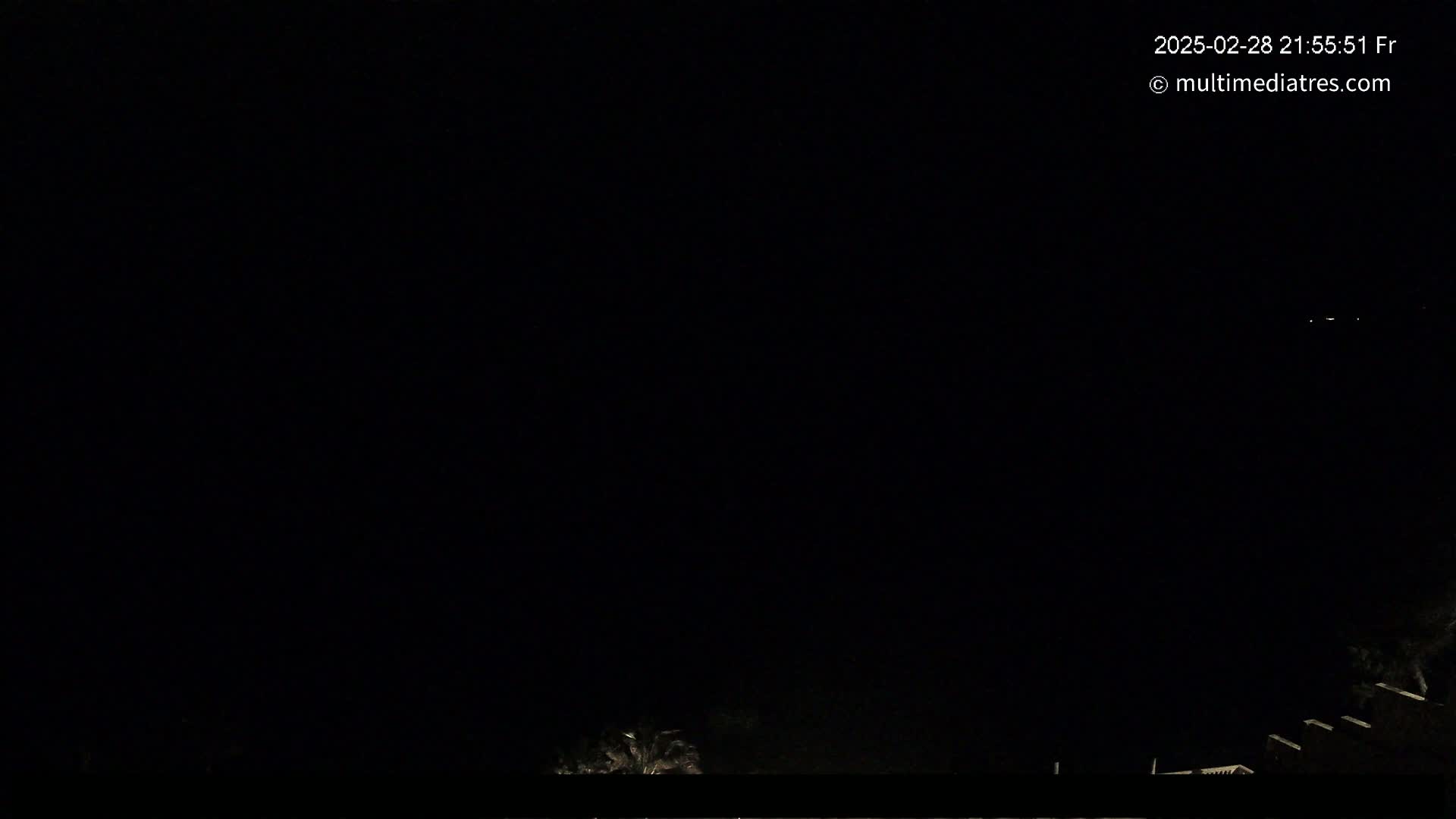 A nighttime outdoor scene shows a very dark sky with a few faint lights in the distance and some indistinct dark shapes, possibly vegetation, in the lower right corner.