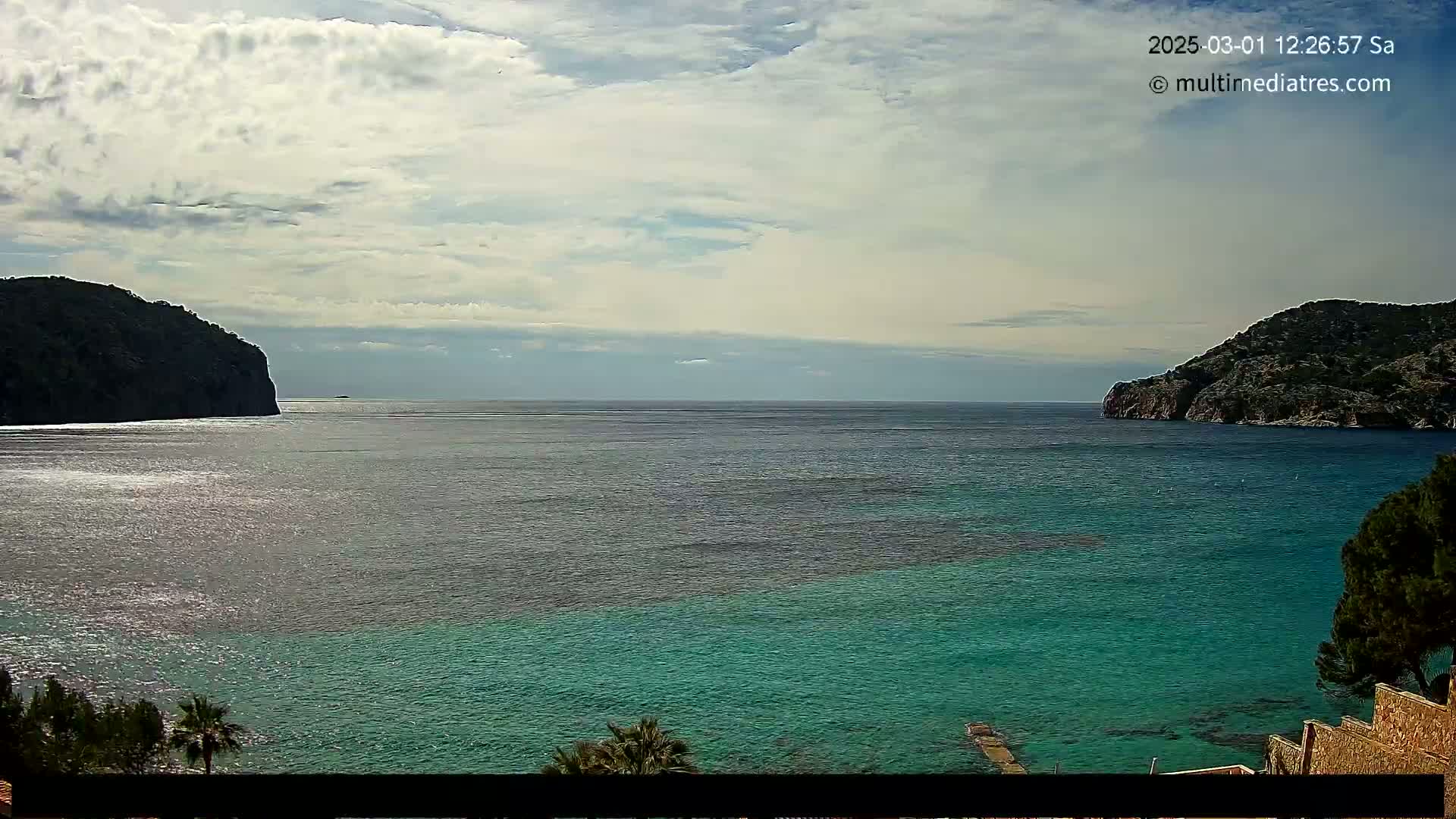 A partly cloudy day shows a tranquil bay of teal water between two dark, rocky hillsides.
