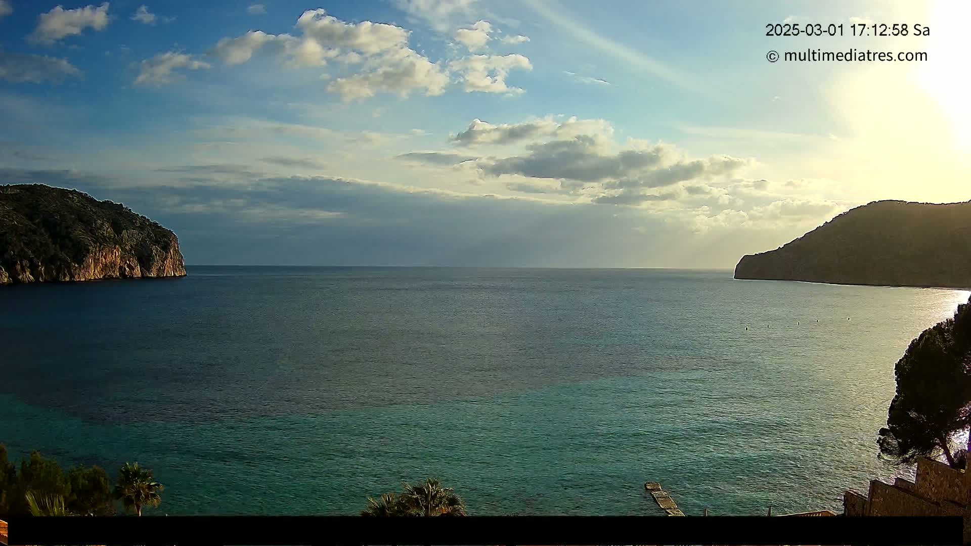 A partly cloudy sky overlooks a calm, teal sea between two dark, rocky headlands.