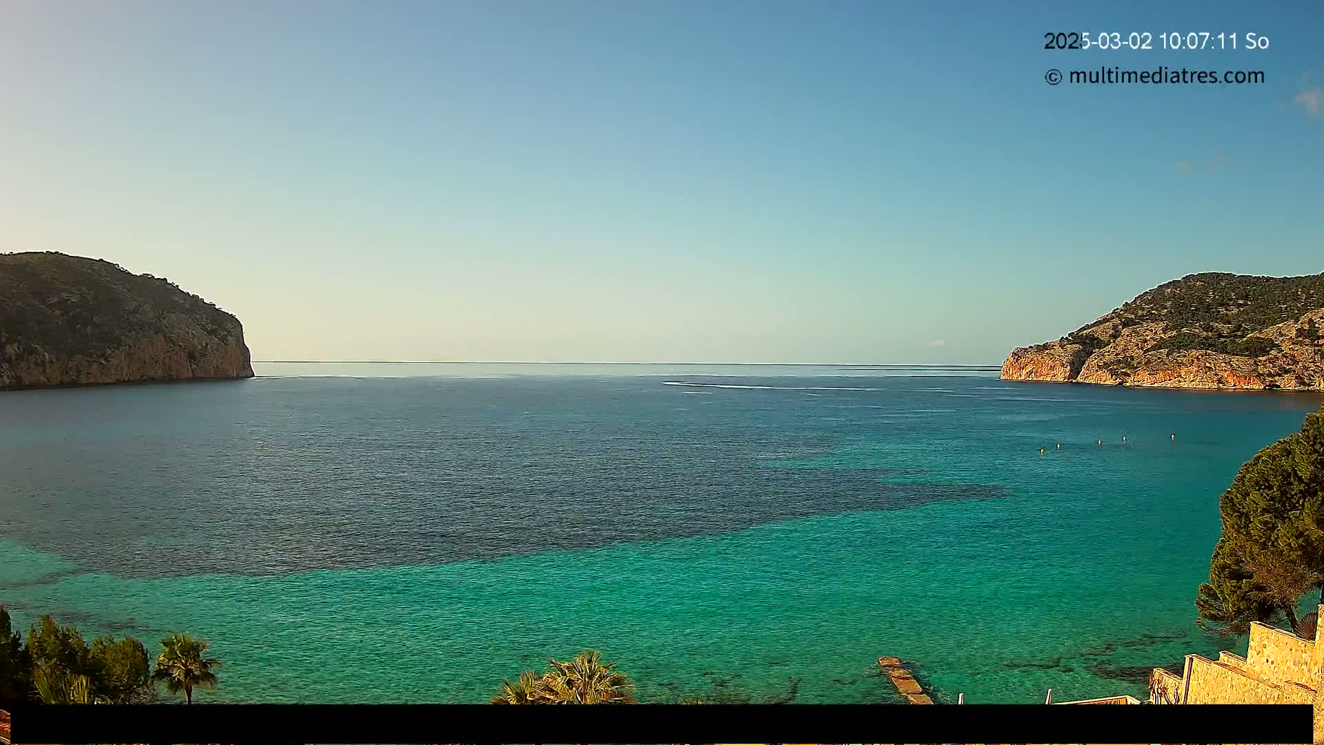A calm, clear day reveals a tranquil bay of vibrant turquoise water nestled between two rocky hillsides.