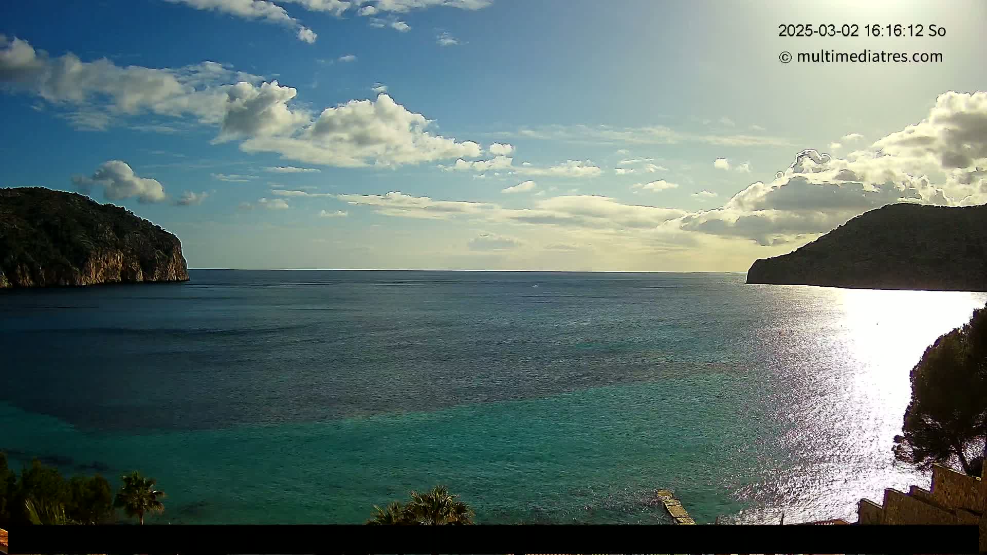 A partly sunny day shows a tranquil bay with dark green water, two rocky hillsides, and some vegetation along the shoreline.