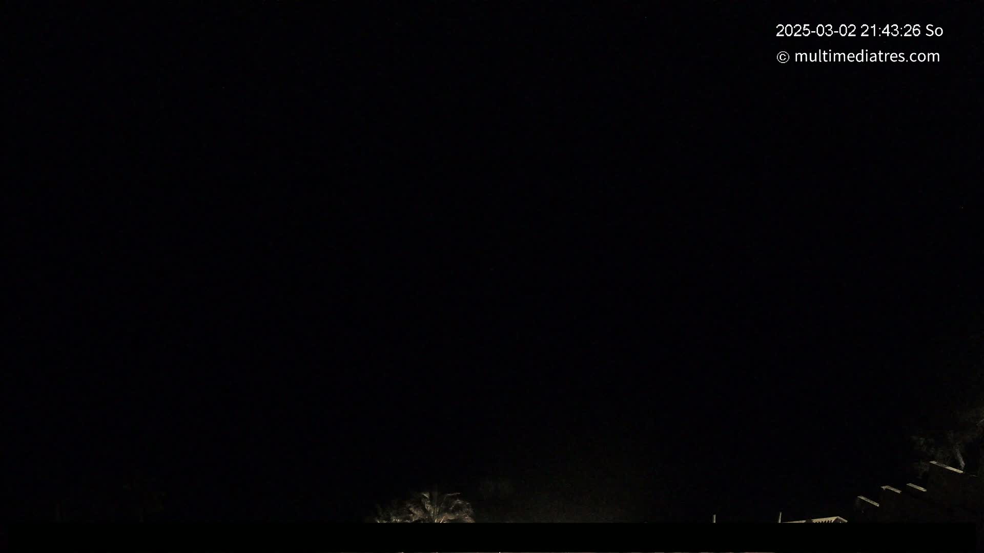 A nighttime, dark outdoor scene shows a few dimly lit trees and structures at the bottom of the frame against a completely black sky.