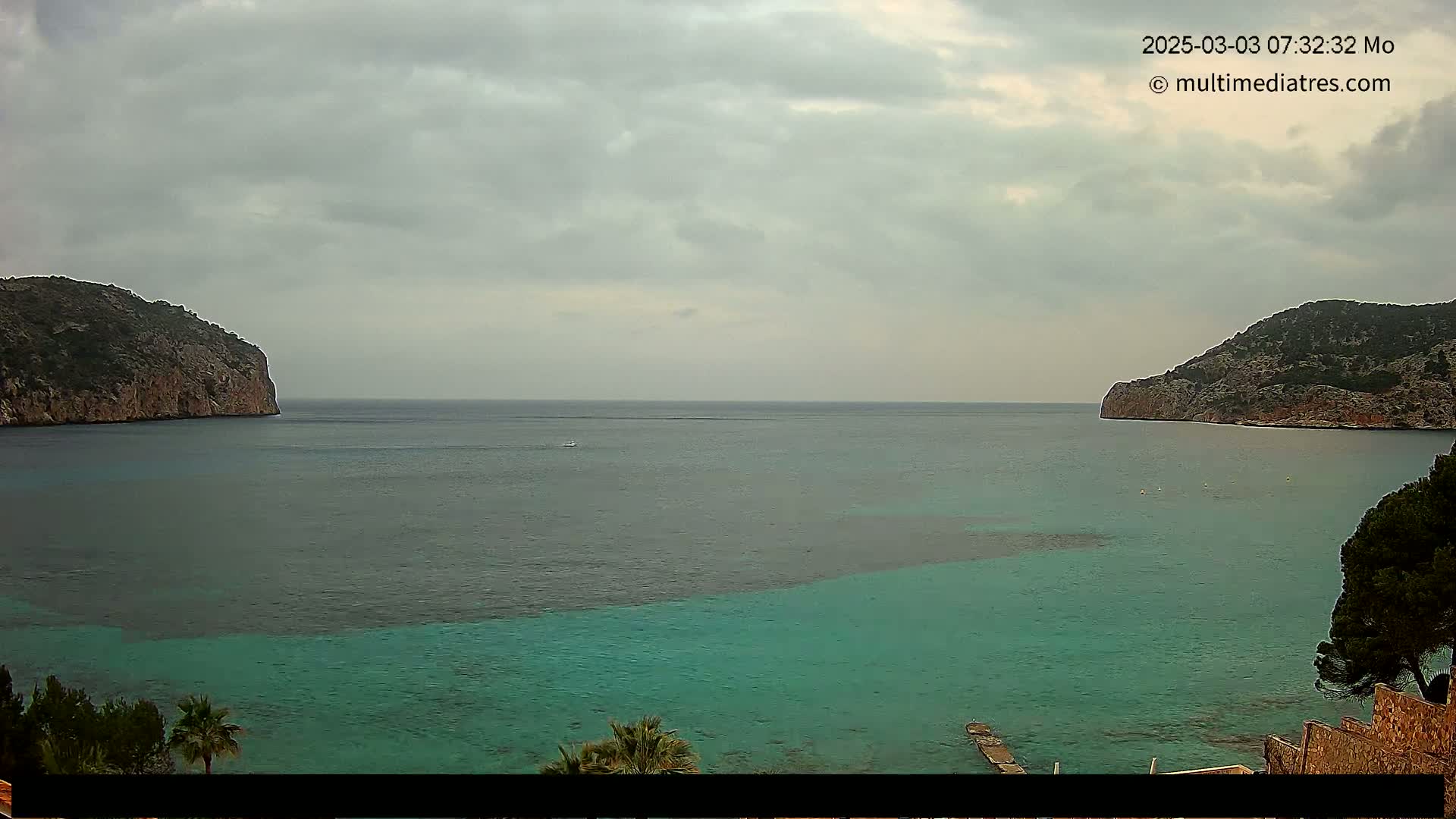 A tranquil bay with calm, teal-colored water is nestled between two rocky hills under a cloudy sky.