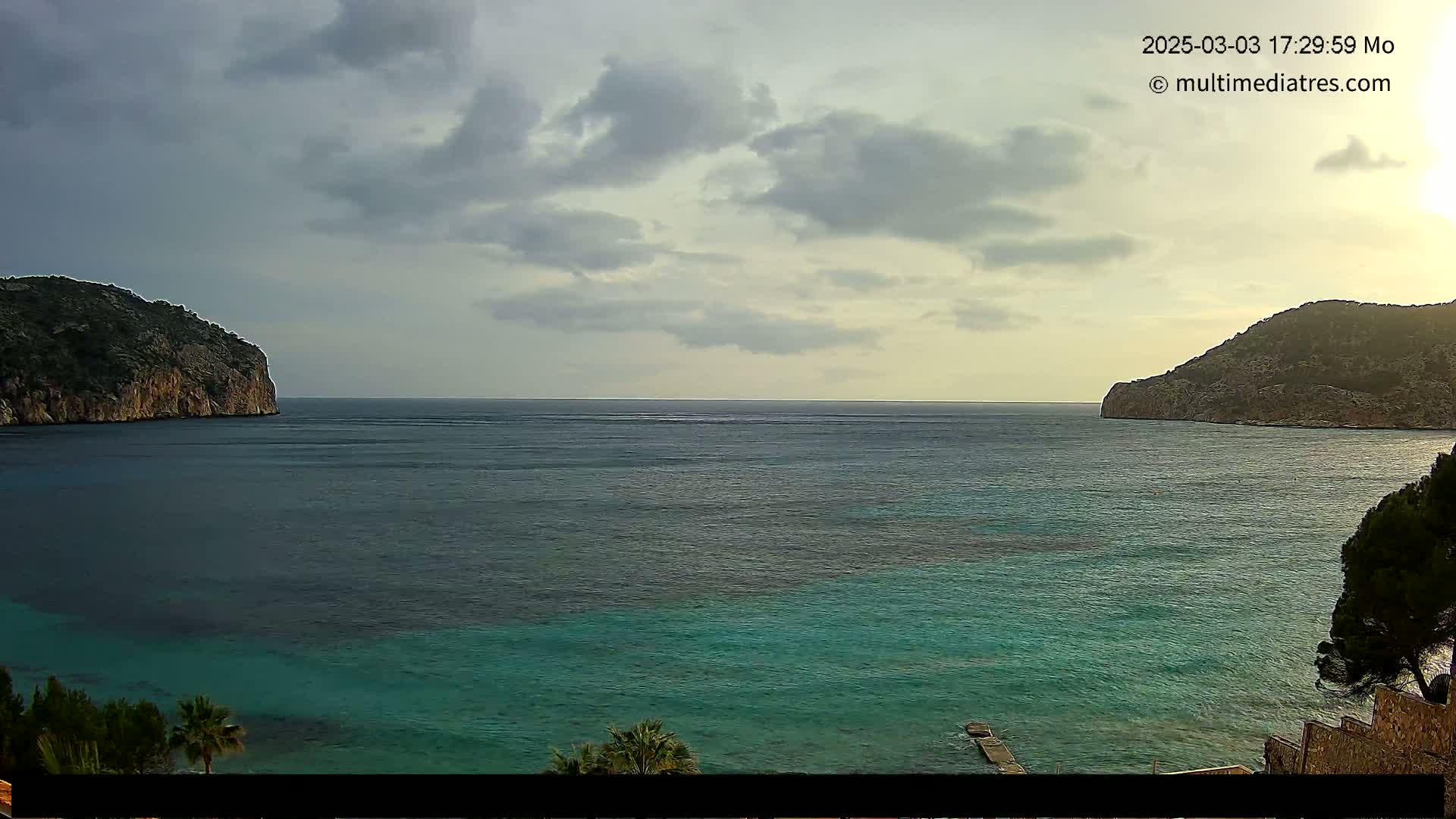A tranquil teal bay is nestled between two rocky hills under a mostly cloudy sky.