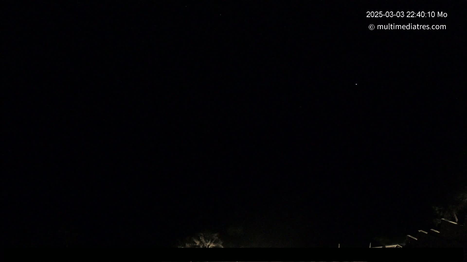 A nighttime outdoor scene shows a mostly clear, dark sky above a low horizon line with indistinct buildings and trees.