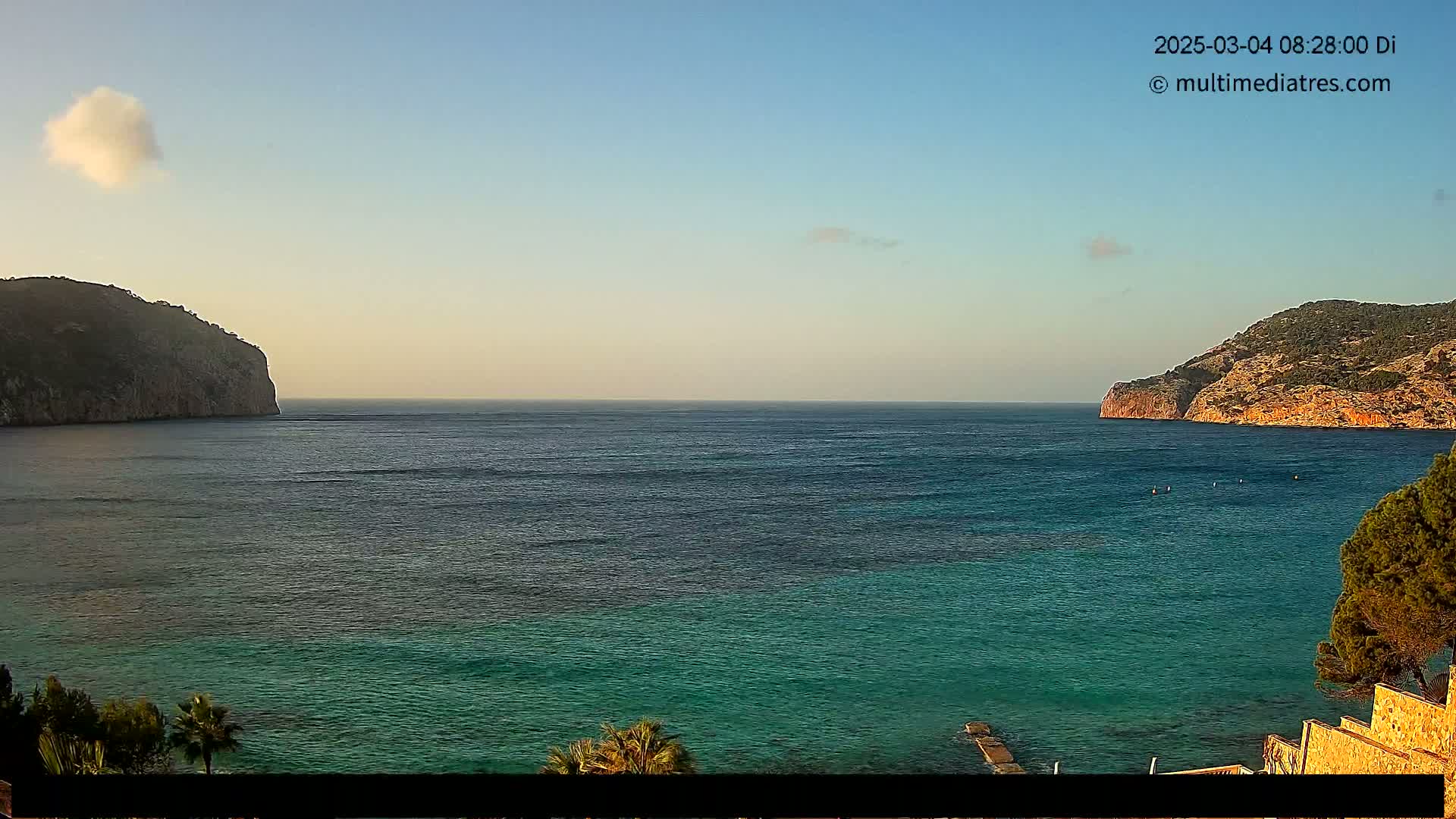 A calm, clear day reveals a tranquil bay with two rocky headlands framing a teal-colored sea.