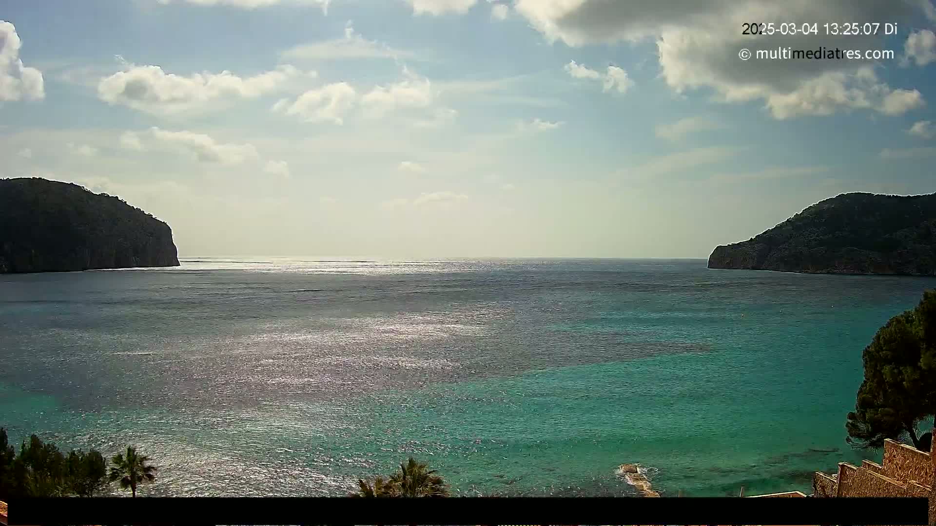 A partly cloudy day reveals a tranquil bay of teal water between two dark, verdant hills.