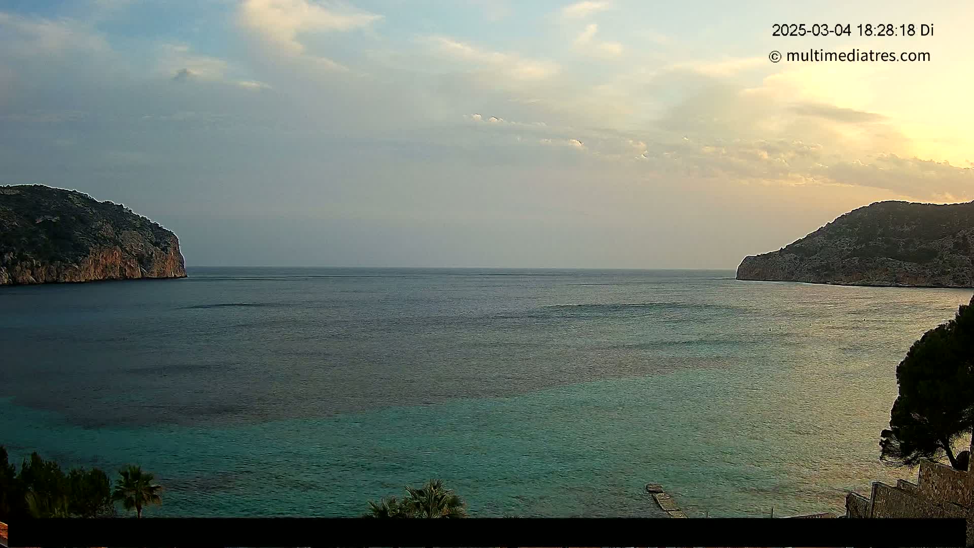 A calm sea with teal-colored shallows near a shoreline, bordered by two rocky hills under a partly cloudy sky.
