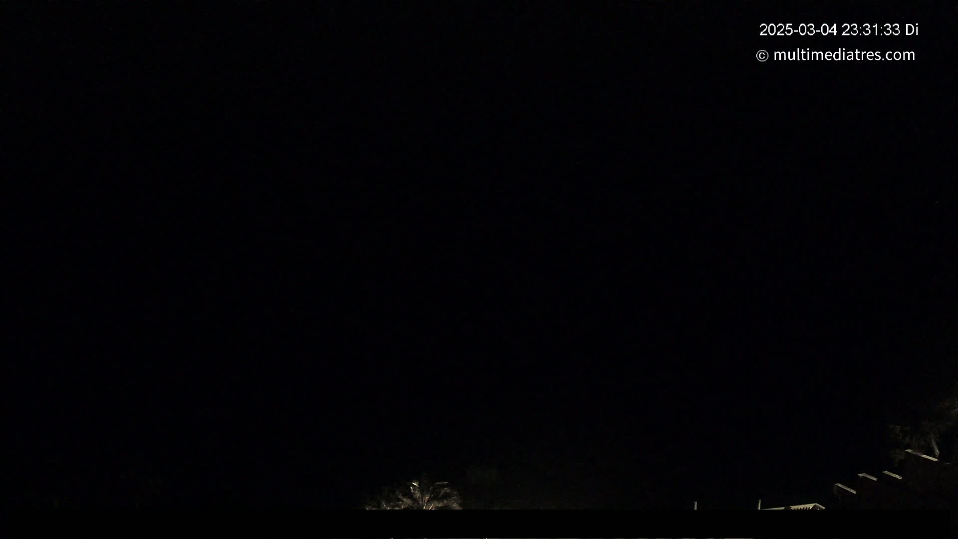 A nighttime, dark scene shows a few dimly lit structures and vegetation at the bottom of the frame against a completely black sky.