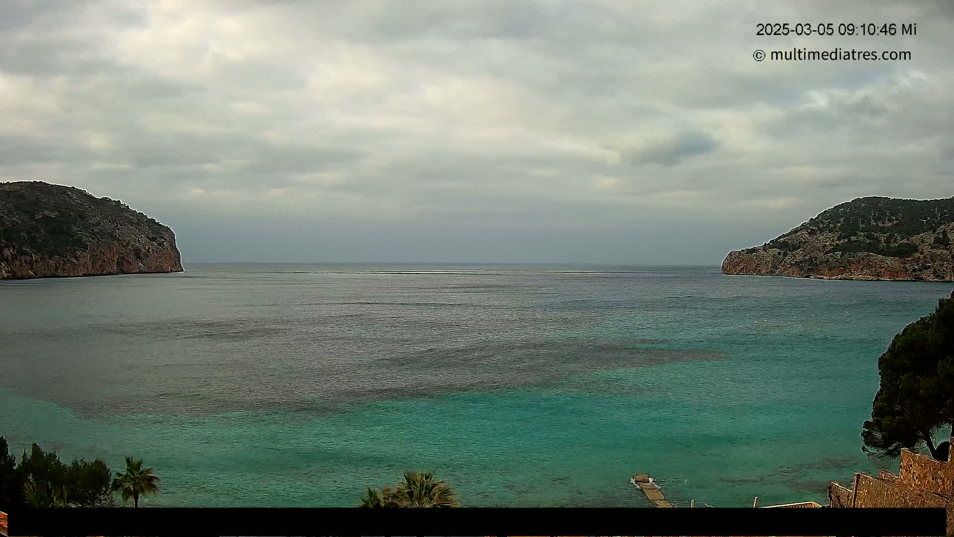 A tranquil bay with teal water is nestled between two rocky hills under a cloudy sky.