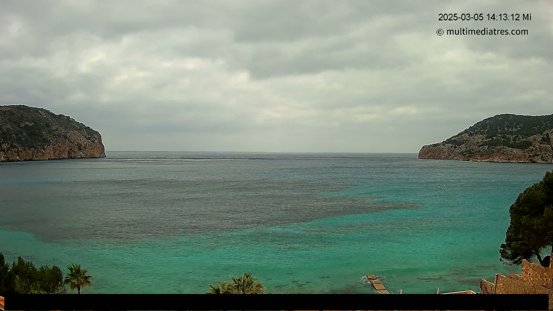 A calm, teal-colored bay is nestled between two rocky hills under an overcast sky.