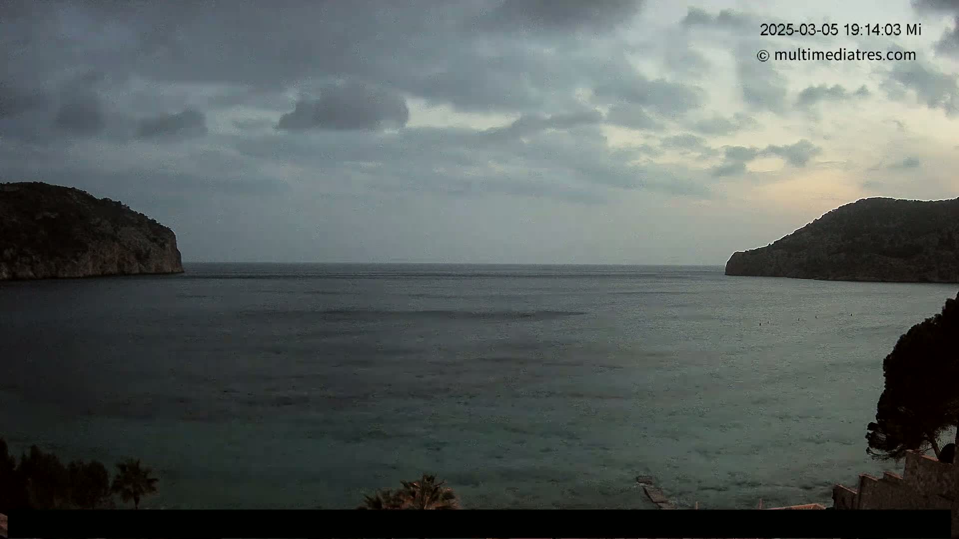 A calm bay is nestled between two dark, rocky hills under a cloudy, overcast sky.