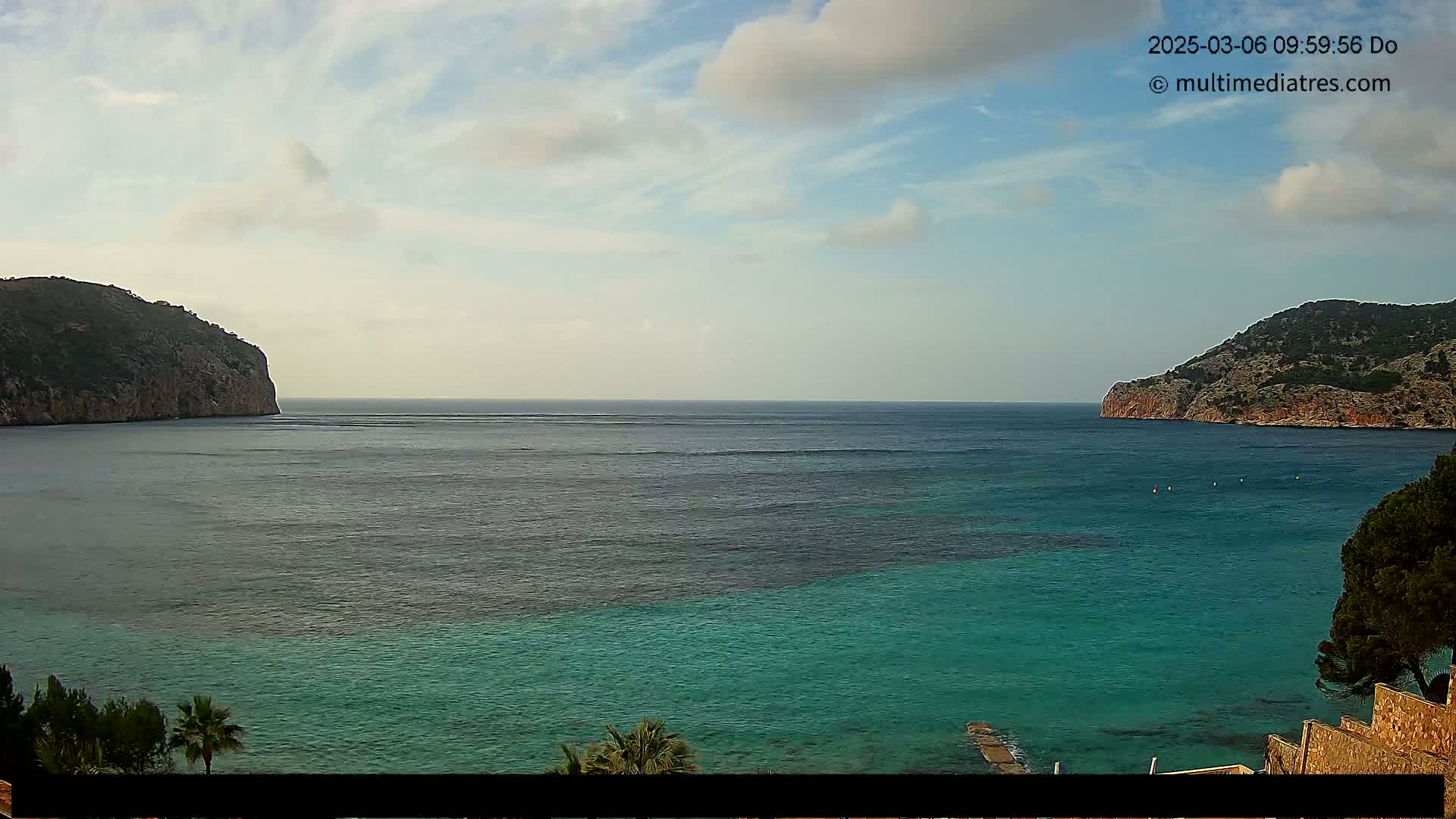 A tranquil cove with clear turquoise water is nestled between two rocky hills under a partly cloudy sky.