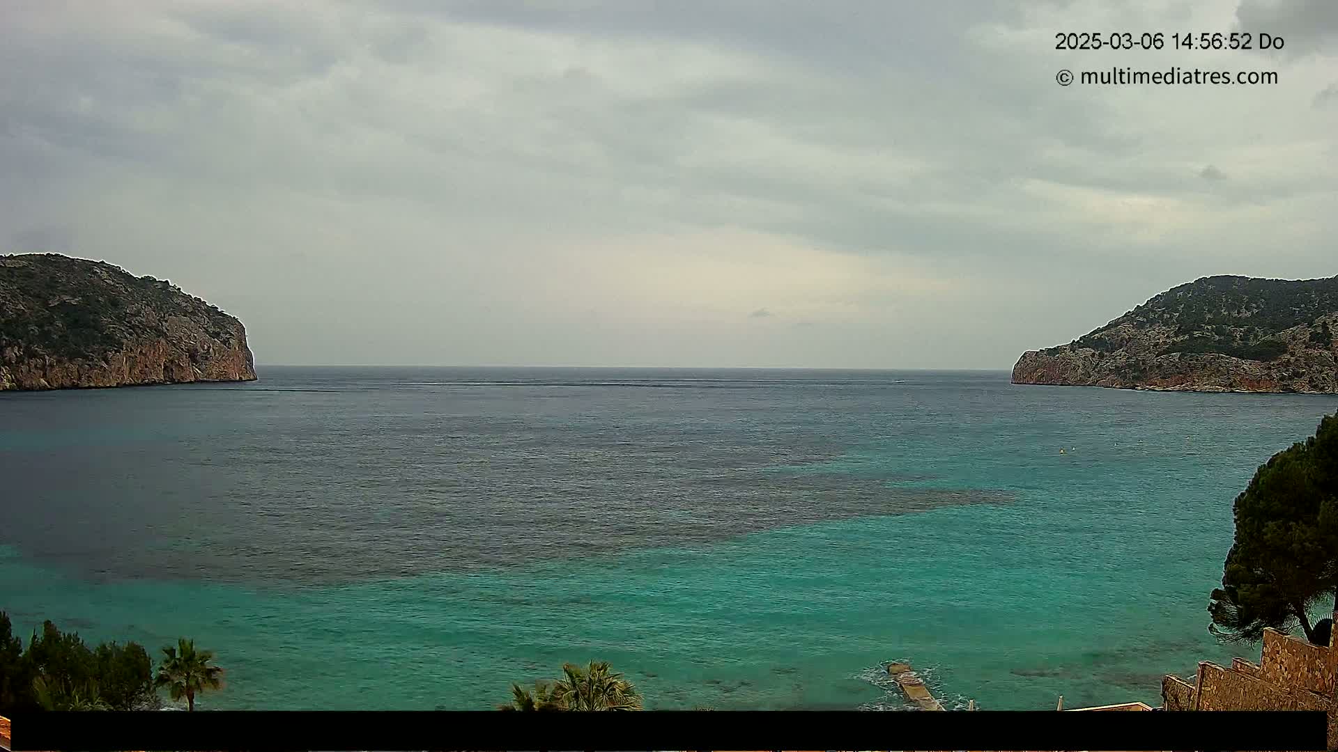 A tranquil cove with teal water is nestled between two rocky hills under an overcast sky.
