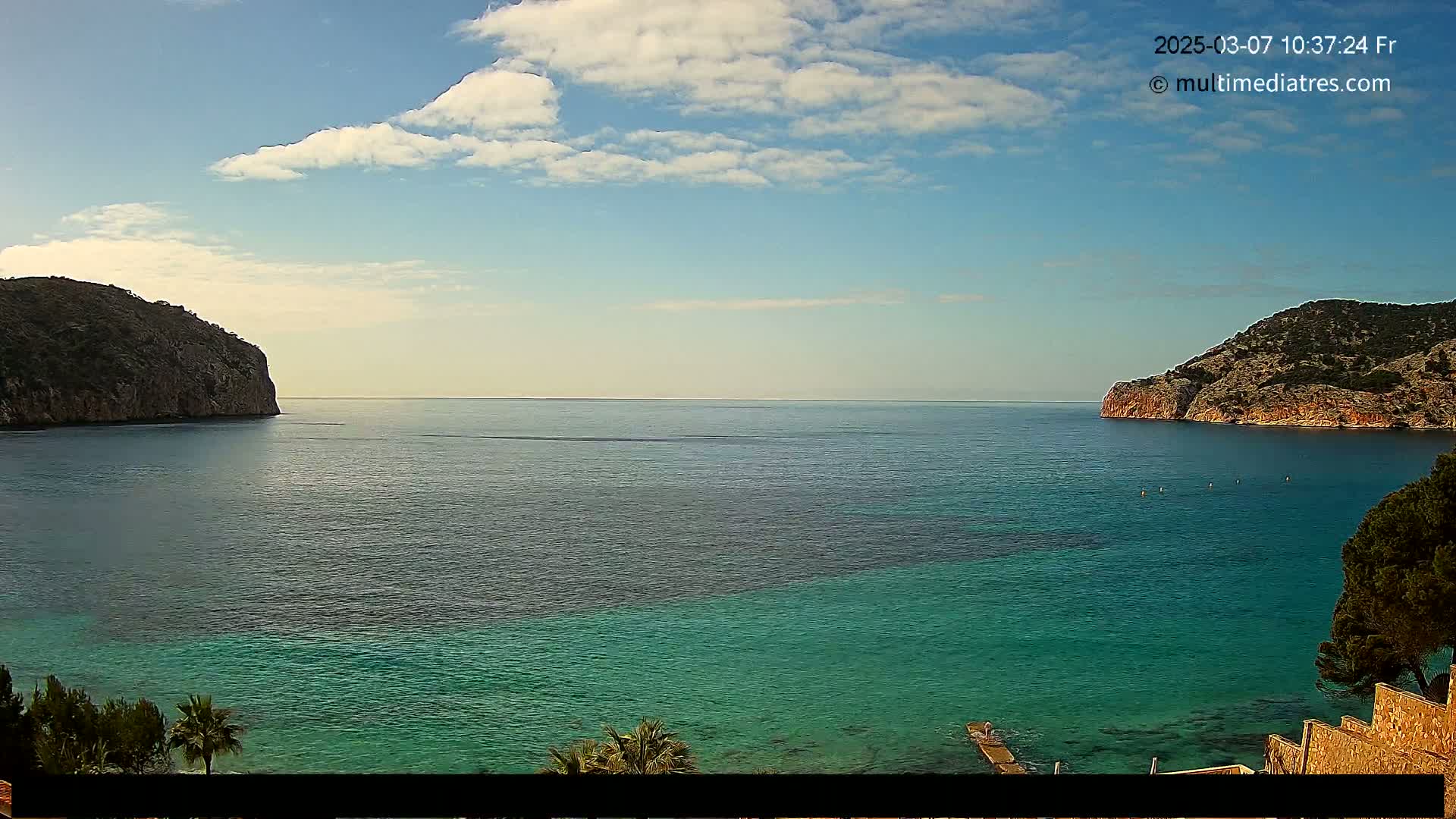 A partly cloudy sky overlooks a calm sea separating two rocky headlands, with the water exhibiting varying shades of blue and green.