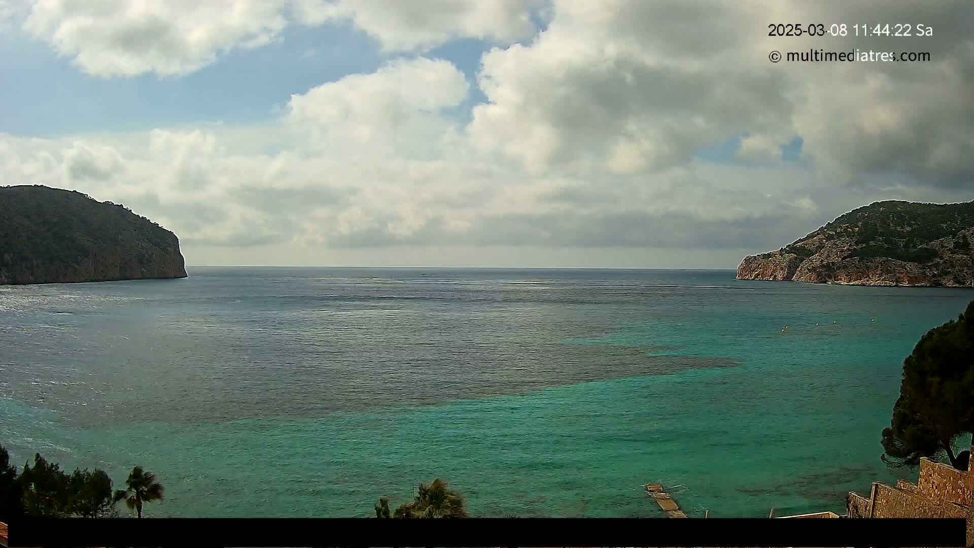 A partly cloudy day reveals a tranquil teal bay nestled between two verdant, rocky hills.