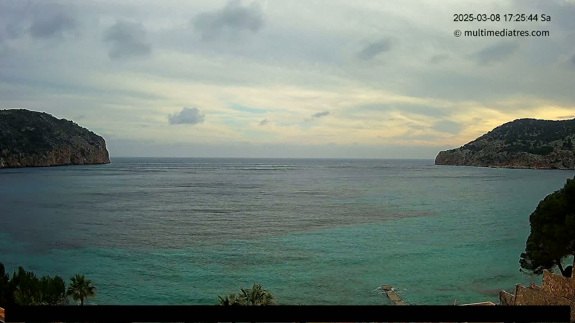 A calm, teal-colored bay is nestled between two rocky, green hills under a cloudy sky.